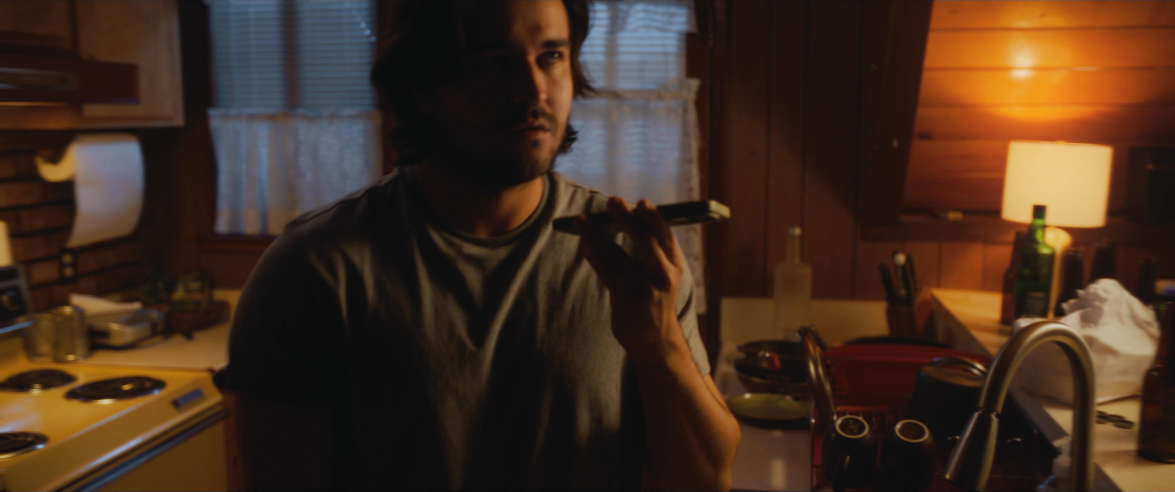 A man speaks on his phone while standing in a kitchen at night, then pauses to open and organize a cluttered utility drawer.