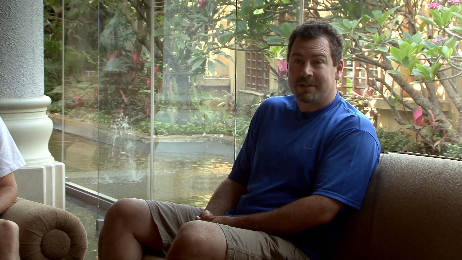 A man in a blue t-shirt sits on a couch while participating in an informal interview or conversation in a well-lit indoor area.