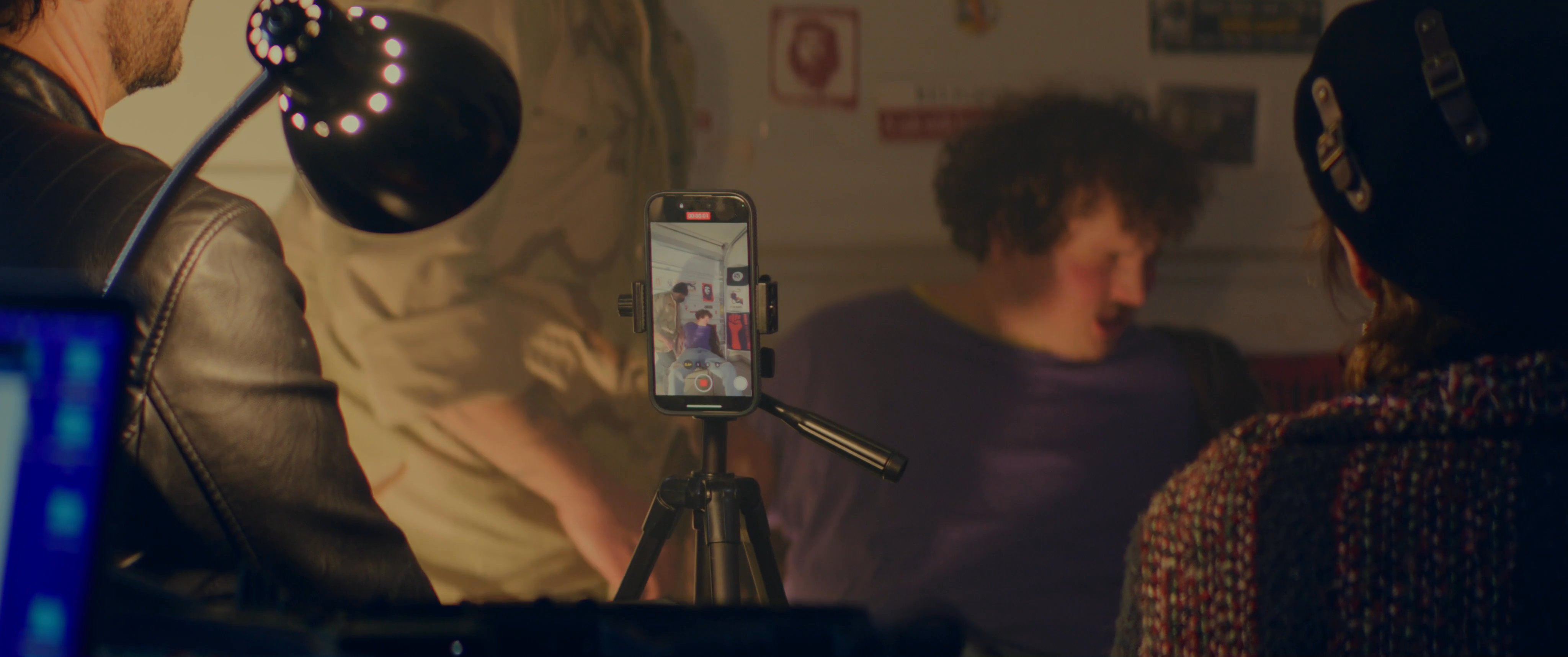 An unidentified man in a mask forcefully compels another person to speak while filming the interaction on a smartphone. A woman observes the scene from behind the camera in a dimly lit, poster-adorned interior.