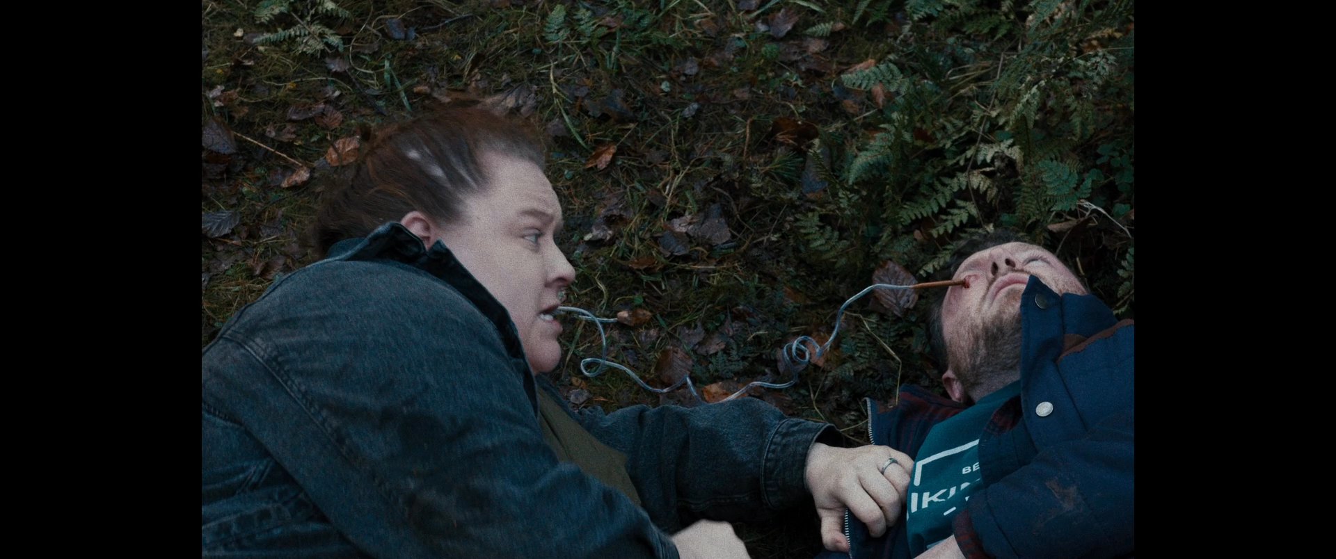 Two individuals engage in a tense physical altercation on the forest floor, with one pinning the other against the earth.