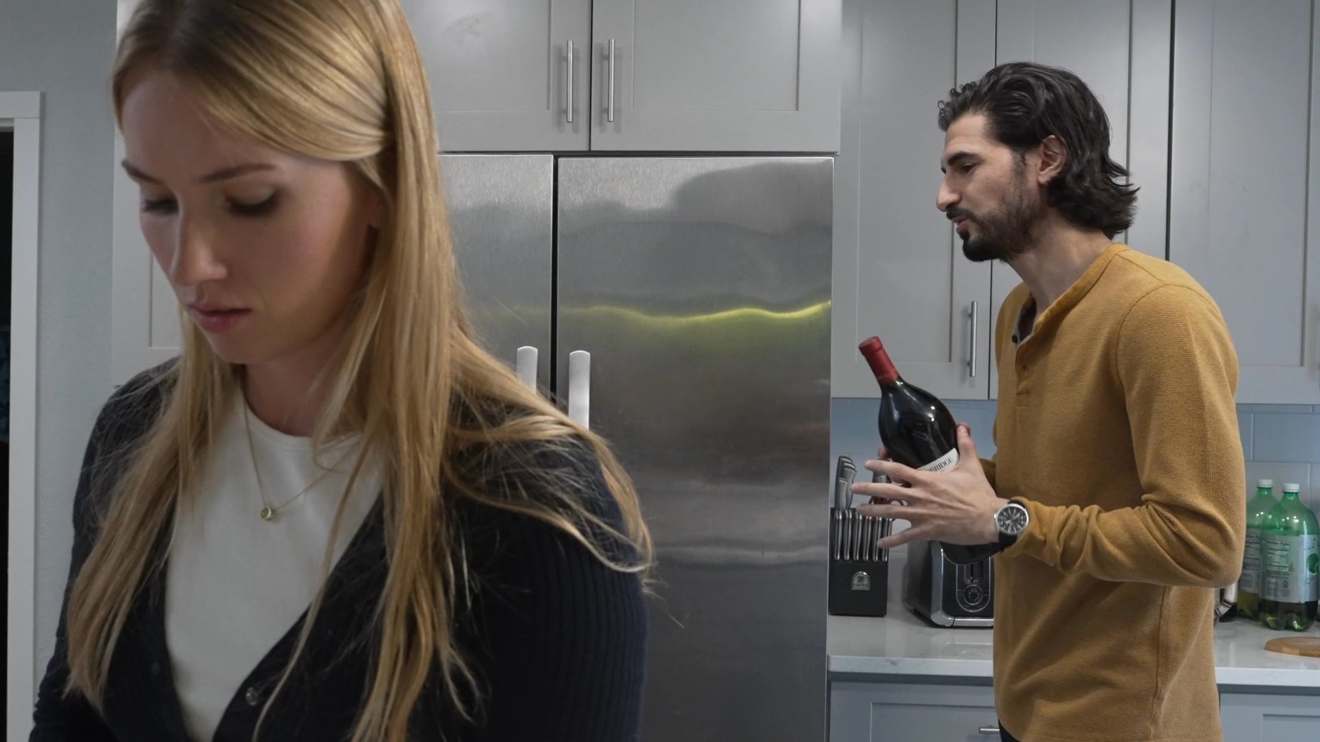 A man holding a wine bottle and a woman holding a kitchen knife engage in a tense, silent confrontation within a residential kitchen setting.