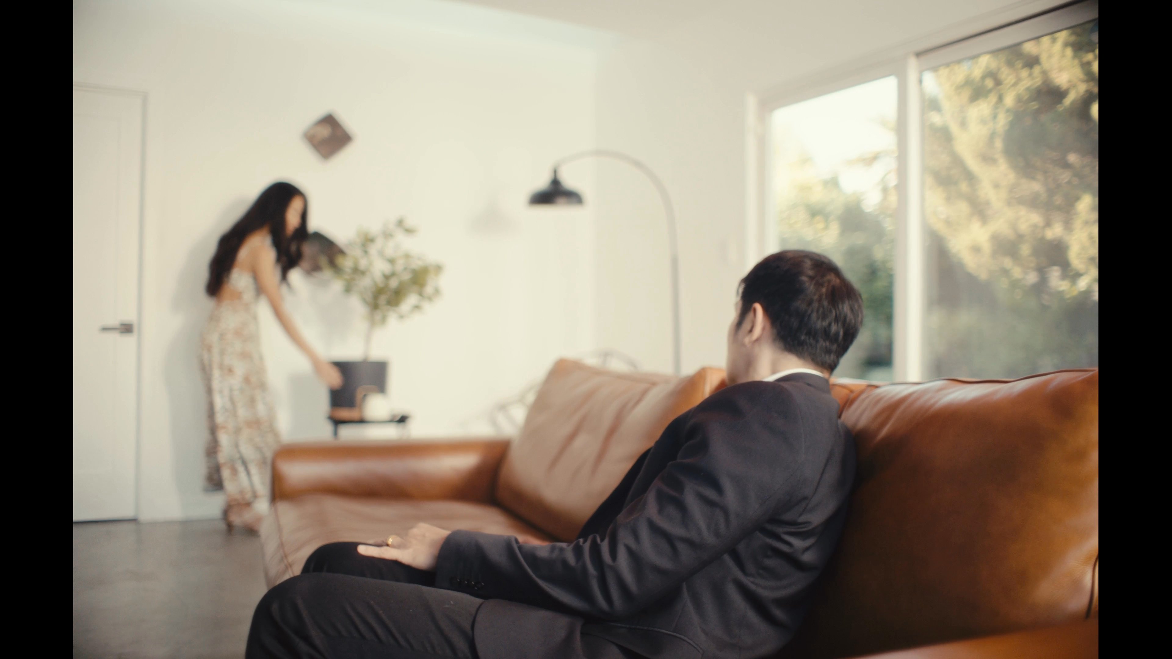 A man wearing a VR headset sits on a sofa, oblivious to his partner who stands apart in the room. The contrast highlights a theme of emotional and physical detachment caused by immersive technology during an intended romantic moment.