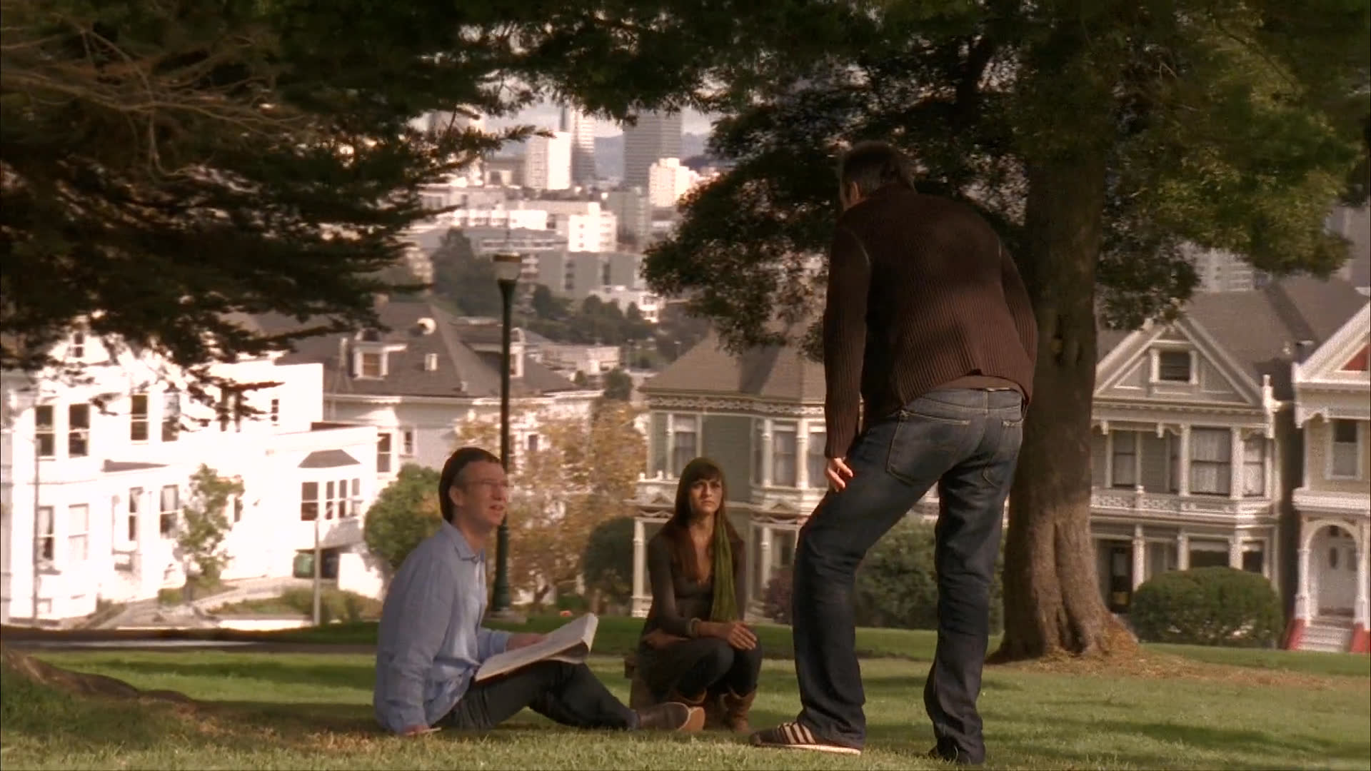A man walks across a grassy park with iconic city architecture in the background to join two people sitting under a tree for a conversation. He engages with them while sharing a small object, initiating a brief social interaction.
