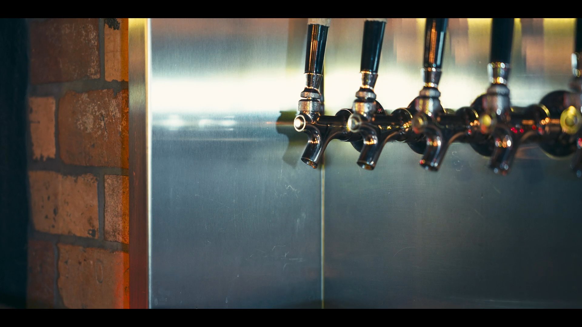 Two patrons sit at a brewery bar counter while a bartender pours a fresh glass of draft beer. The atmosphere is casual as the guests converse and relax in the industrial-style setting.