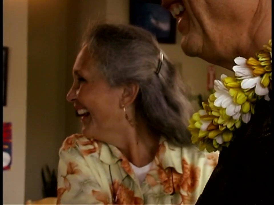 Marilyn Burns engages in warm social interactions with guests at a reception, where participants are adorned with traditional flower leis.
