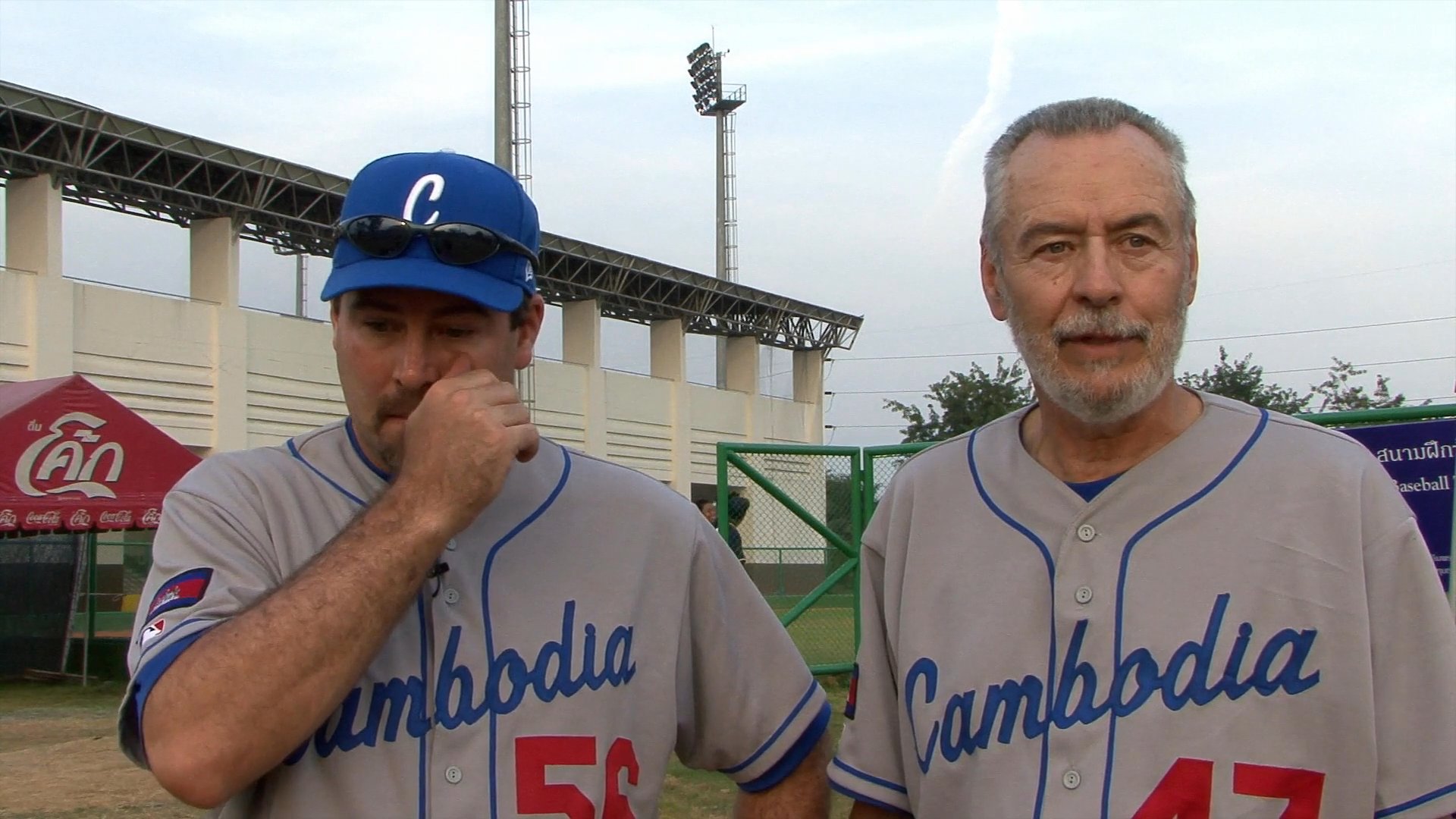Two men in Cambodia baseball team jerseys provide commentary following a game against the Philippines at a stadium venue.