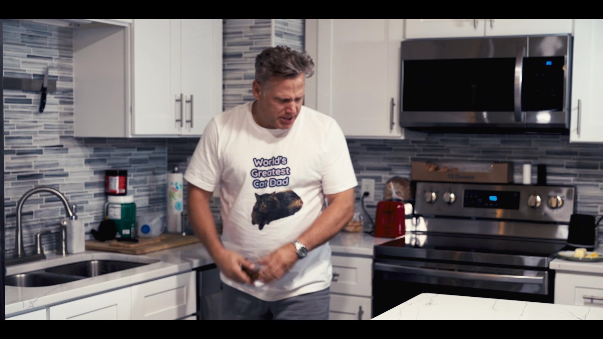 A man pours liquid from a container into a mug within a modern kitchen setting. He concludes by adjusting his shirt and observing the completed drink.