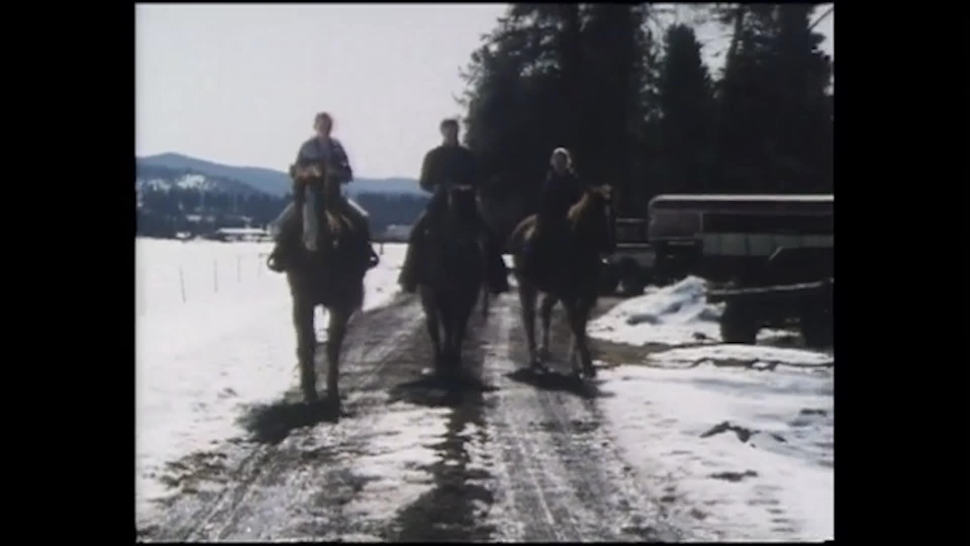 A young girl in a purple jacket smiles beside a wooden wall as a group of riders approaches along a muddy, snow-lined road. They follow directions to turn and continue through gates near the forest.