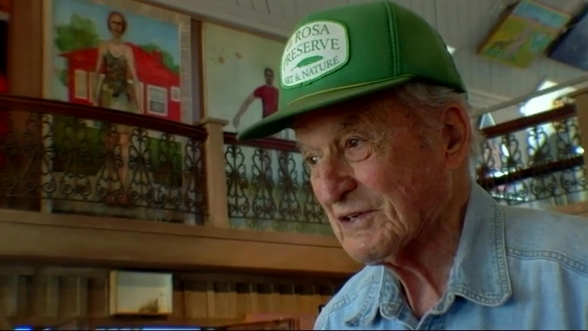An older man in a green cap speaks inside an art-filled gallery, reflecting on the pleasure of discovering work by artists who have never sold their art. The sequence ends with another man seated among artworks, handling a large sheet or portfolio.