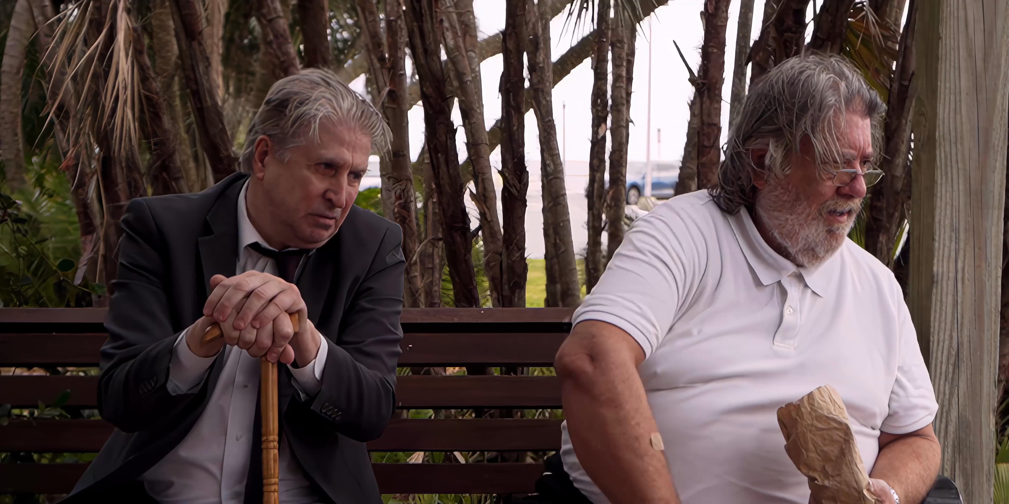Two men engage in a thoughtful conversation while seated on a park bench surrounded by foliage. The dialogue appears focused and intimate as they exchange perspectives in an outdoor setting.