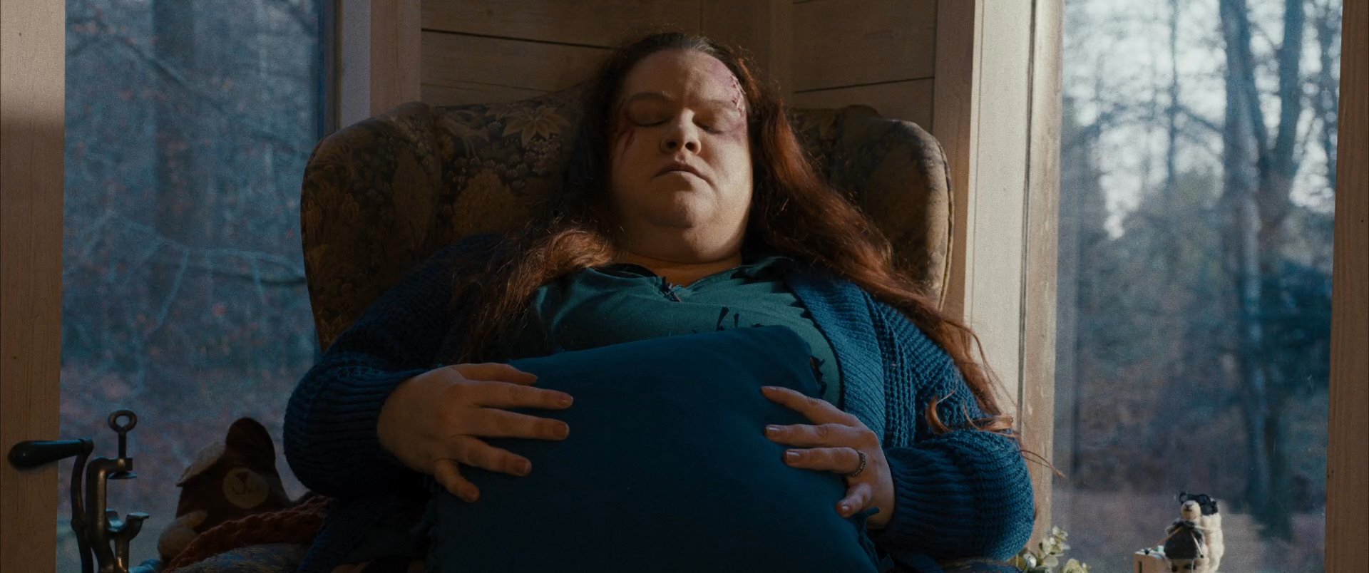 A woman sits in an armchair by a window, gently resting her hands on her stomach while lost in deep thought.