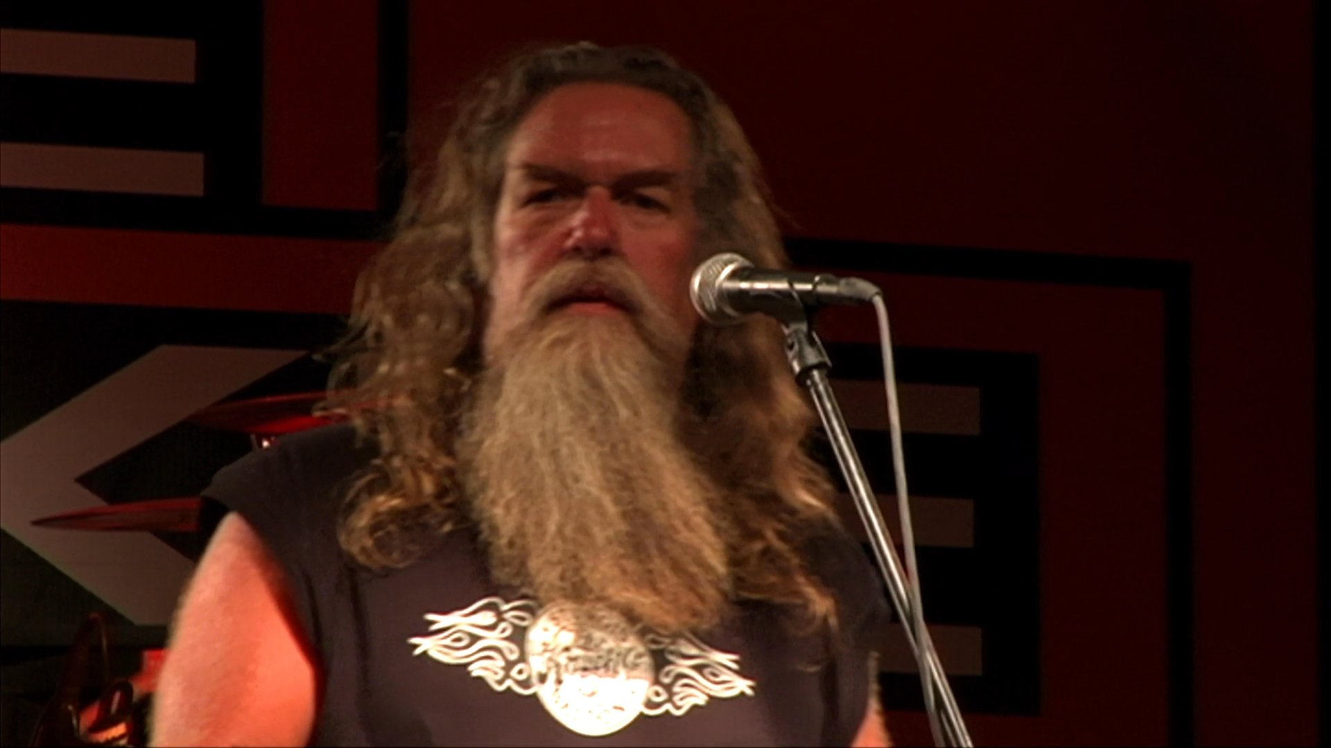 A long-haired male singer performs on stage with his band at a music venue. The footage transitions to nostalgic, grainy archival film showing individuals carrying a casket at a funeral.