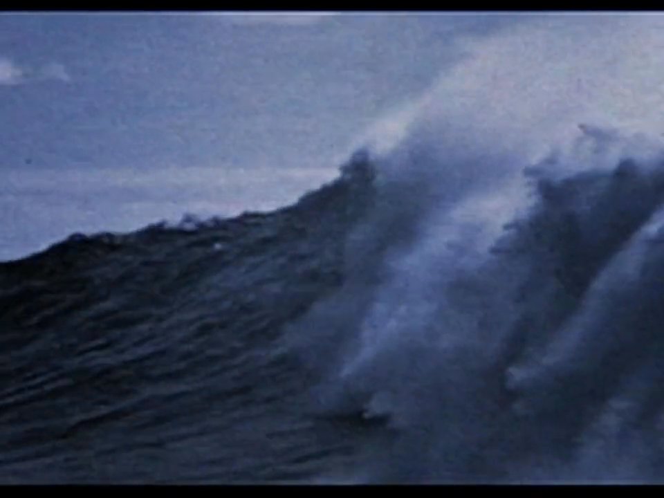 A surfer navigates a massive ocean wave, demonstrating expert control and skill as the water breaks along a coastal environment.