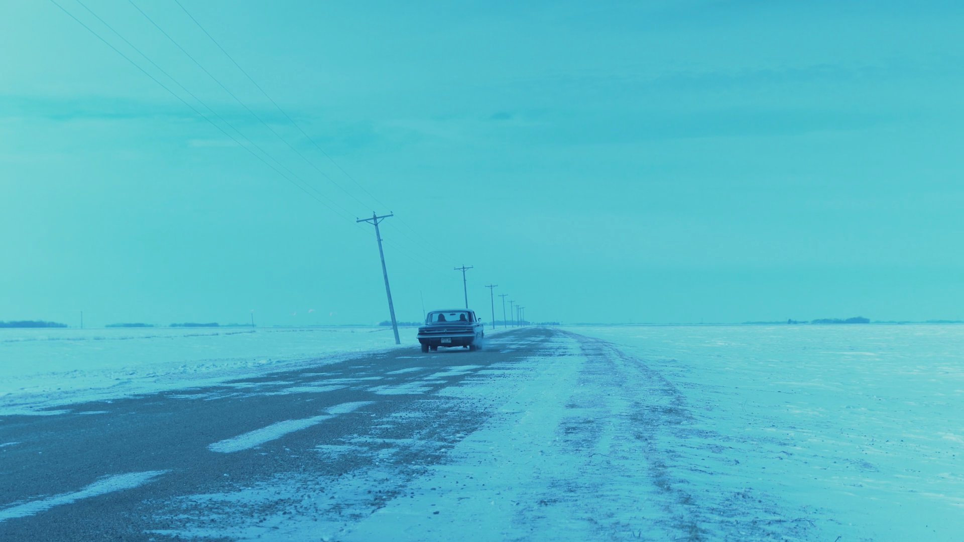 A dark sedan travels along a desolate, snow-covered rural road. The sequence concludes with a shot of a man and woman driving inside the vehicle.
