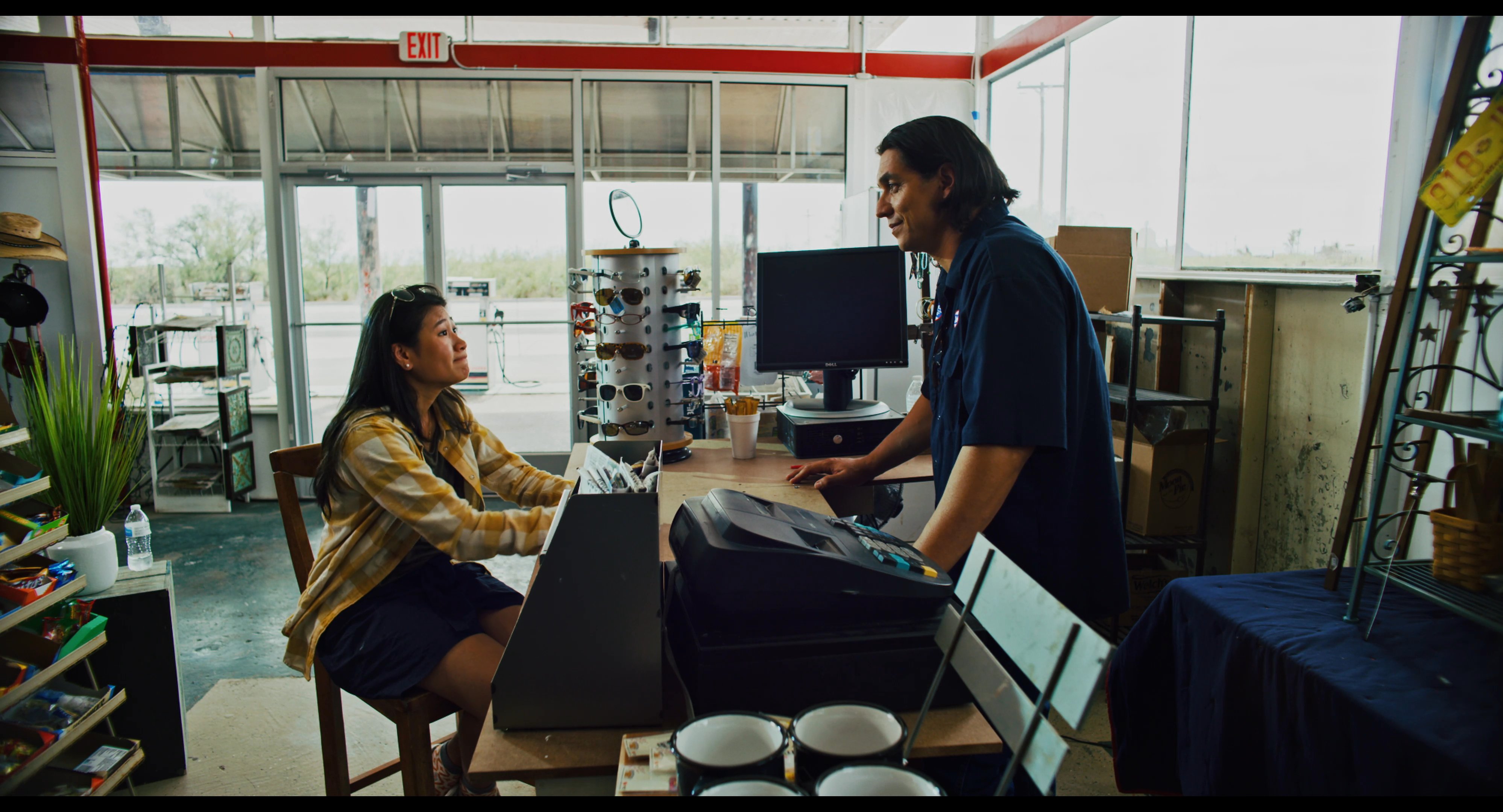 A man in a service uniform discusses security matters with a woman inside a retail establishment. The scene concludes with a focus on a security camera warning sign displayed on the glass door.
