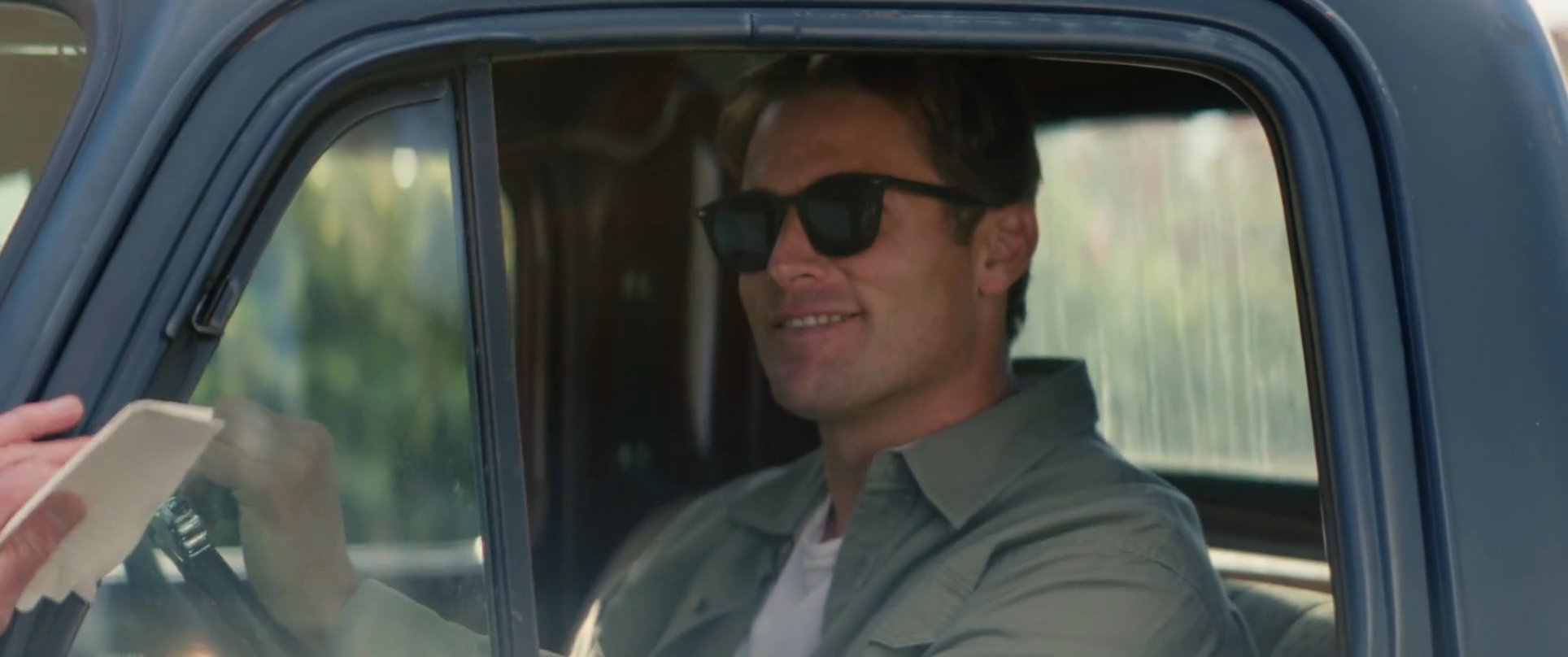 Two men exchange a handshake before one departs in a classic blue pickup truck, driving away from a suburban residence.