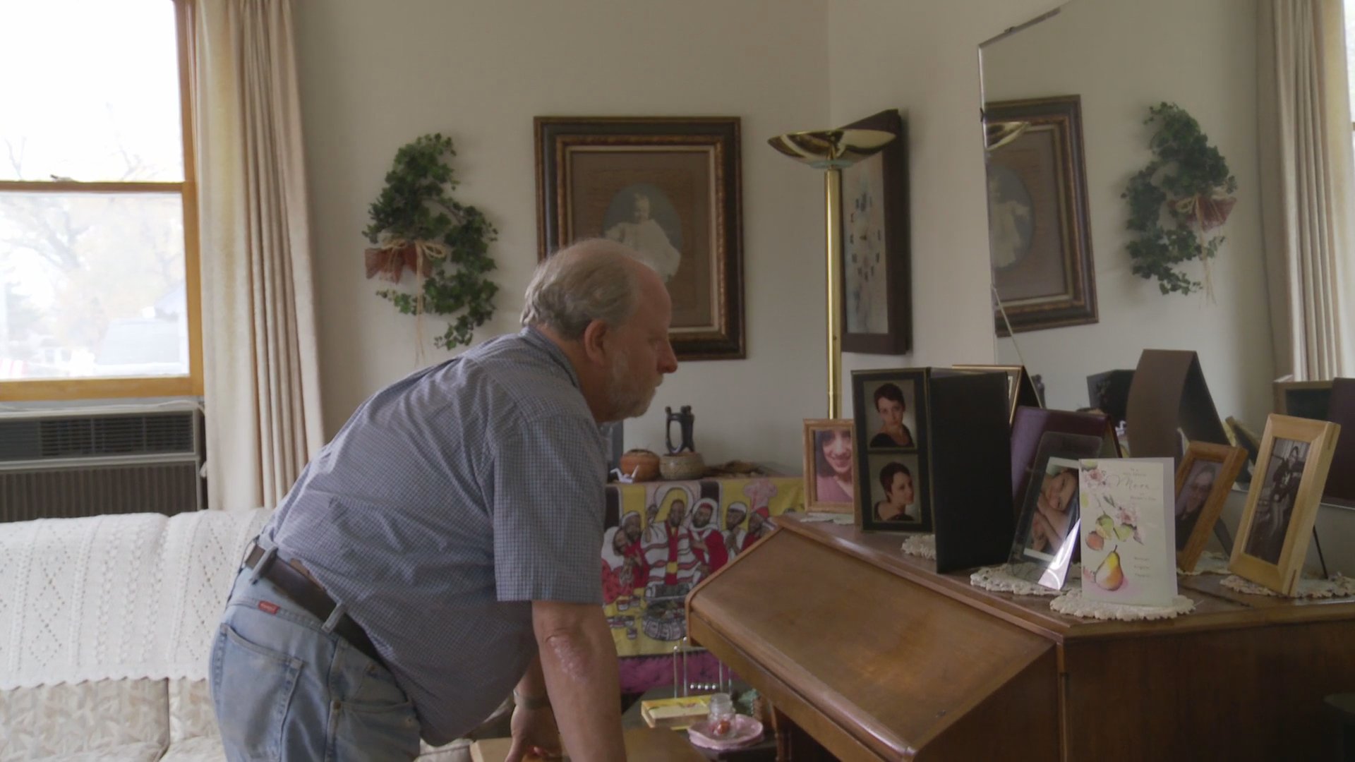 An elderly woman engages in an intense phone conversation while seated in her living room, as her partner moves about the home in the background. The scene captures a moment of domestic tension and emotional exchange between the two individuals.