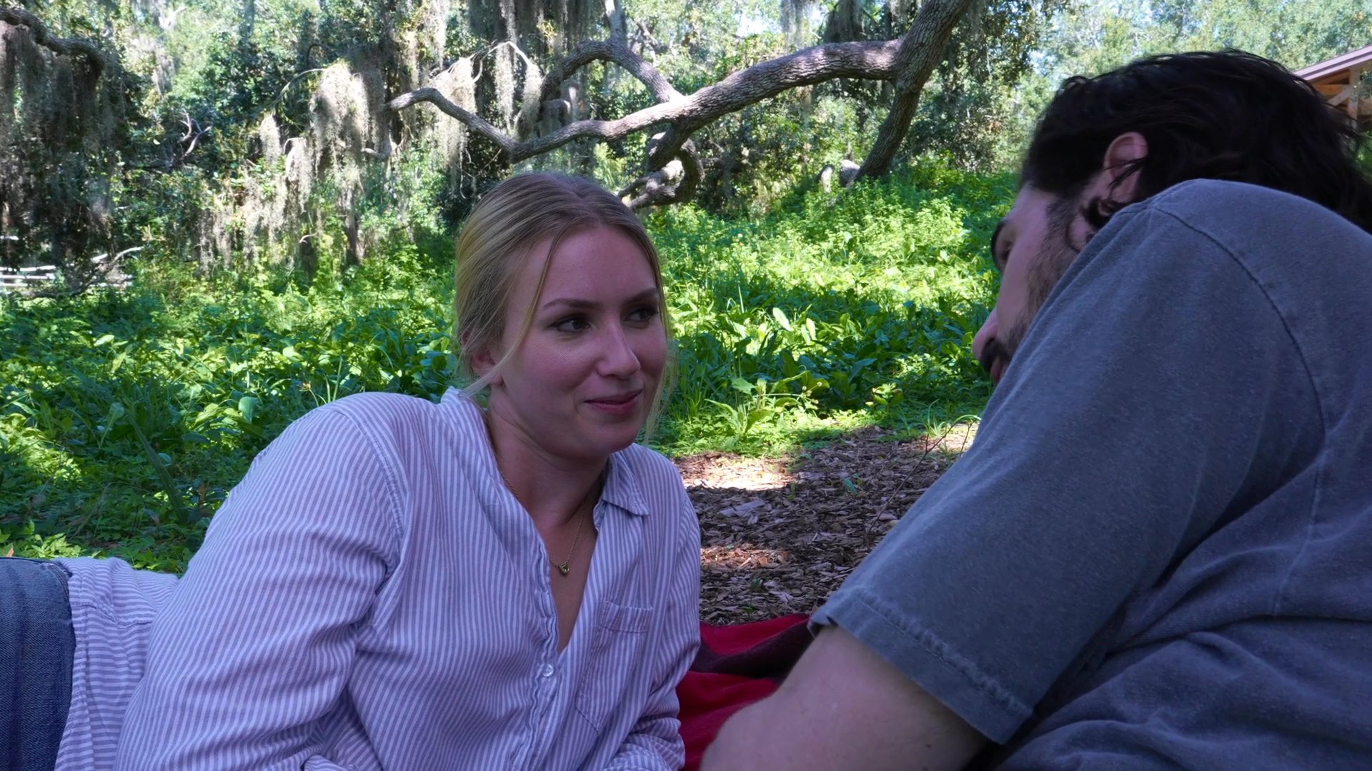 A man and a woman lie on a blanket in a park, engaging in a lighthearted conversation while resting in a wooded, sunlit area.