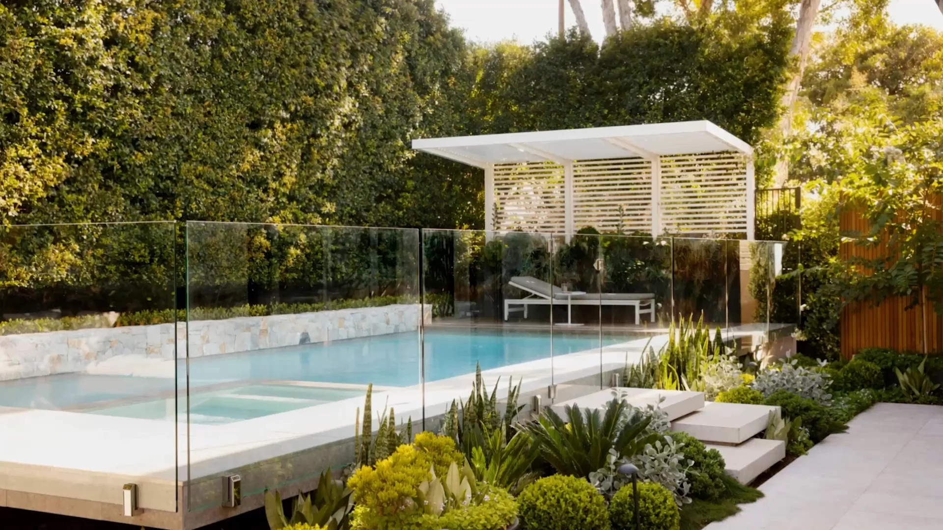 Floating pool and spa behind glass fencing with white slatted pergola and layered native garden at dusk