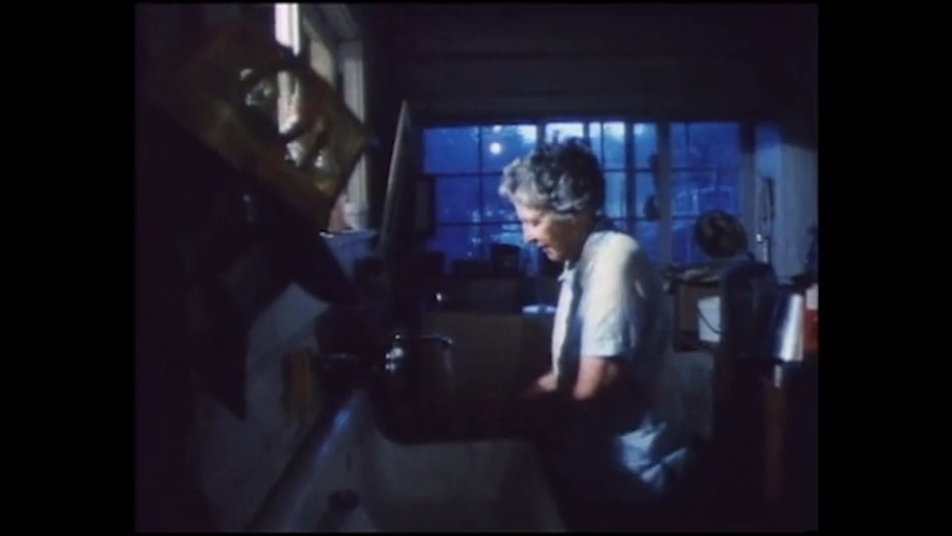 A man works at the stove and prepares food at a kitchen counter before an older woman tends to tasks near the sink. She bends over her work and then looks up with a warm smile as the narration recalls Norma’s practical skills and outdoor nature.