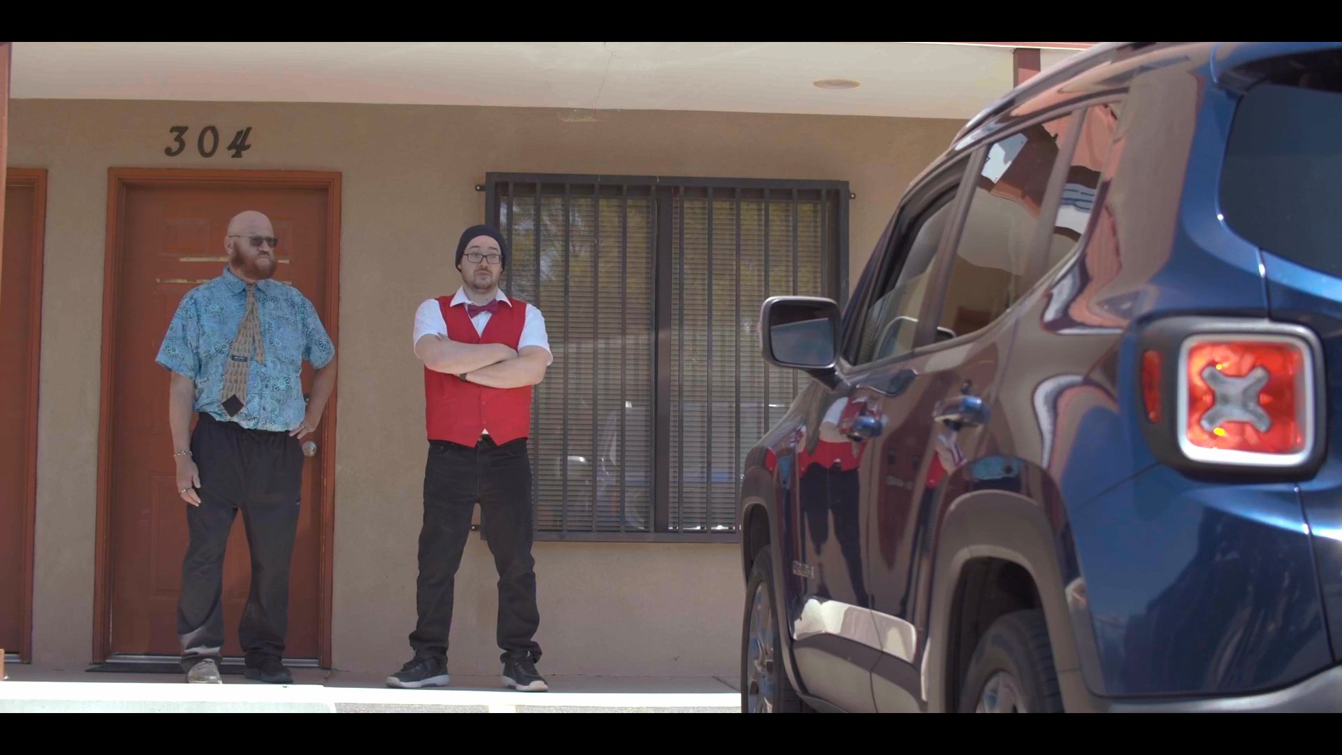 Individuals exchange items before departing in a vehicle parked outside a motel unit. The scene establishes a professional yet enigmatic interaction between the involved parties.