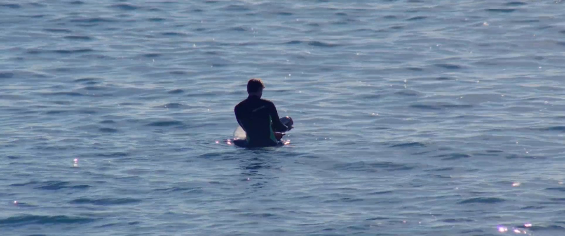 An individual in a wetsuit shares an emotional embrace on a sandy beach before entering the ocean to paddle out into the surf.