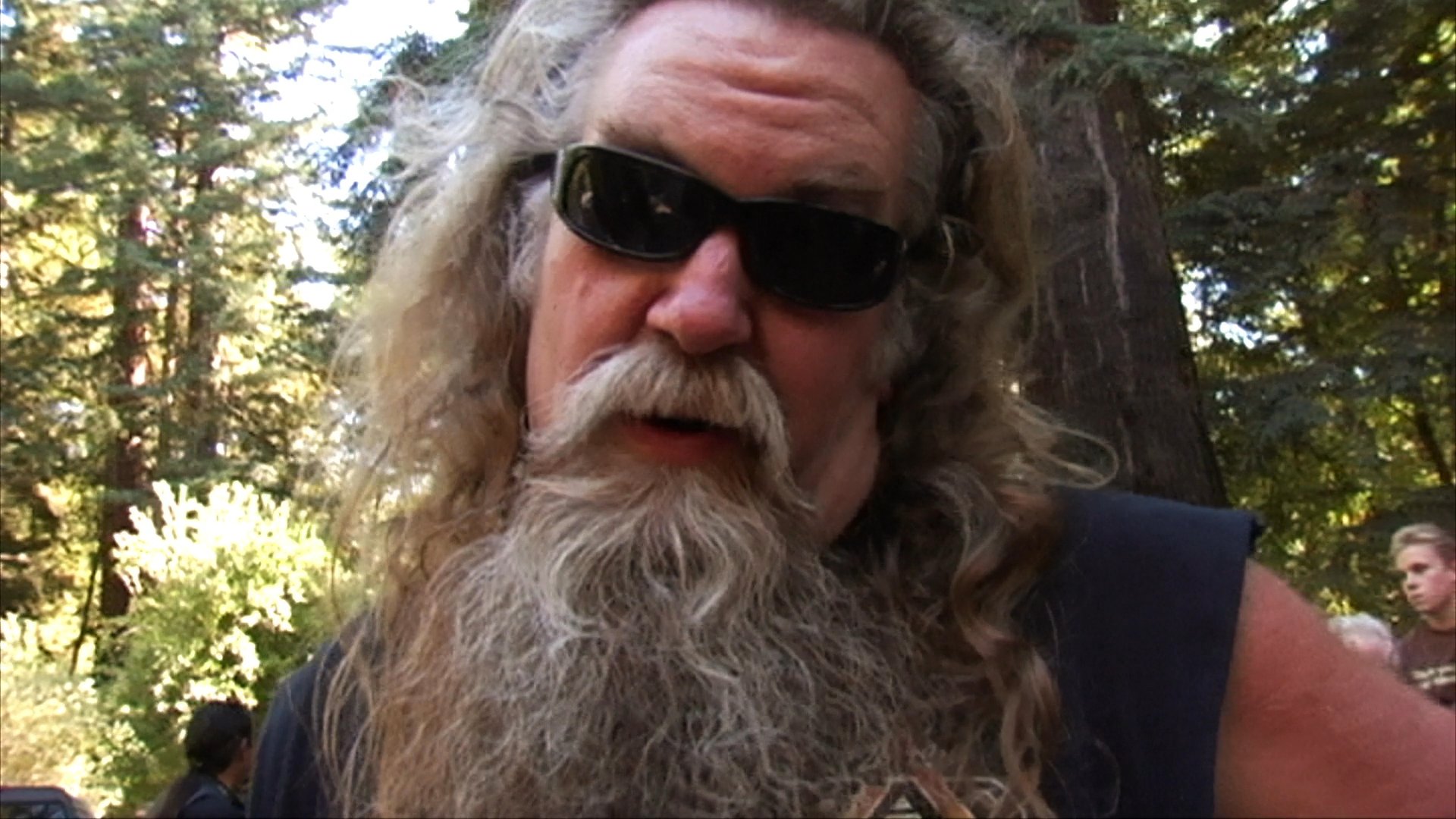 Two bearded men in biker attire discuss community experiences while situated in a wooded outdoor environment.