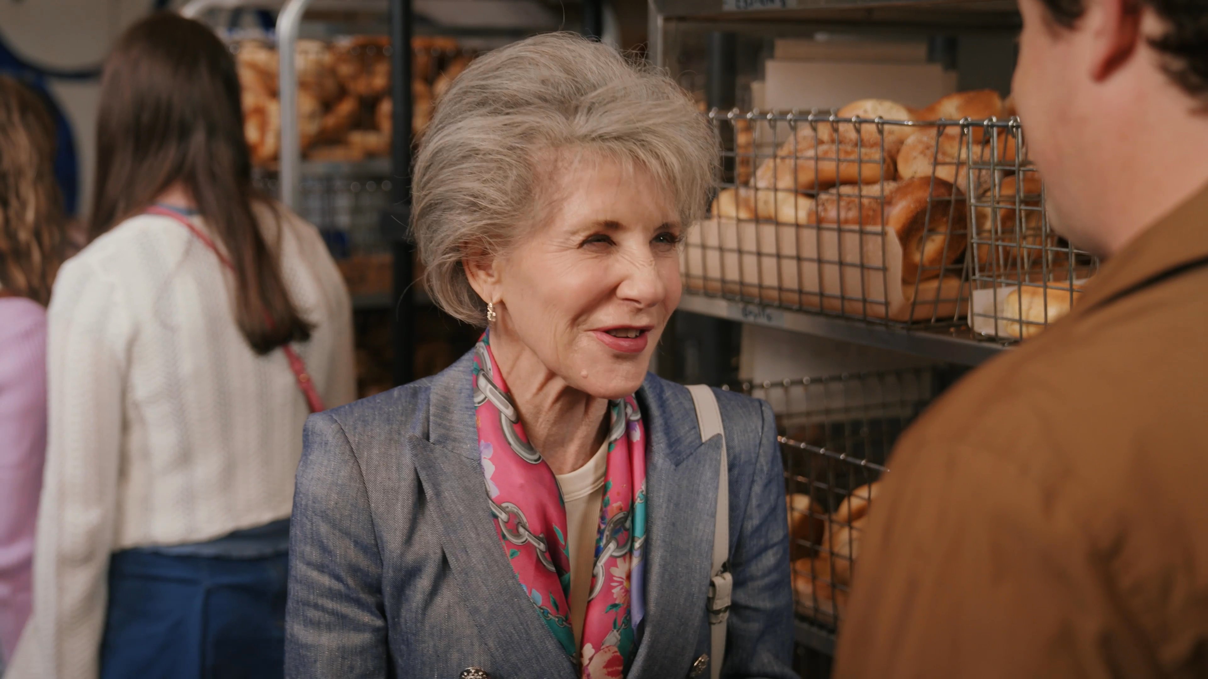 An older woman engages in a pleasant conversation with a younger man while standing in a busy bakery shop. They exchange words near the bread display shelves as other customers wait in the background.