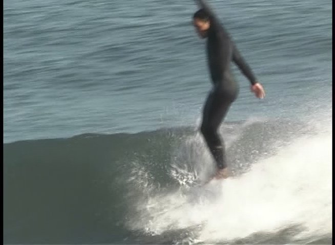 A surfer wearing a full-body wetsuit navigates a breaking wave while maintaining balance on a surfboard. The athlete expertly maneuvers across the face of the wave along a coastal shore.