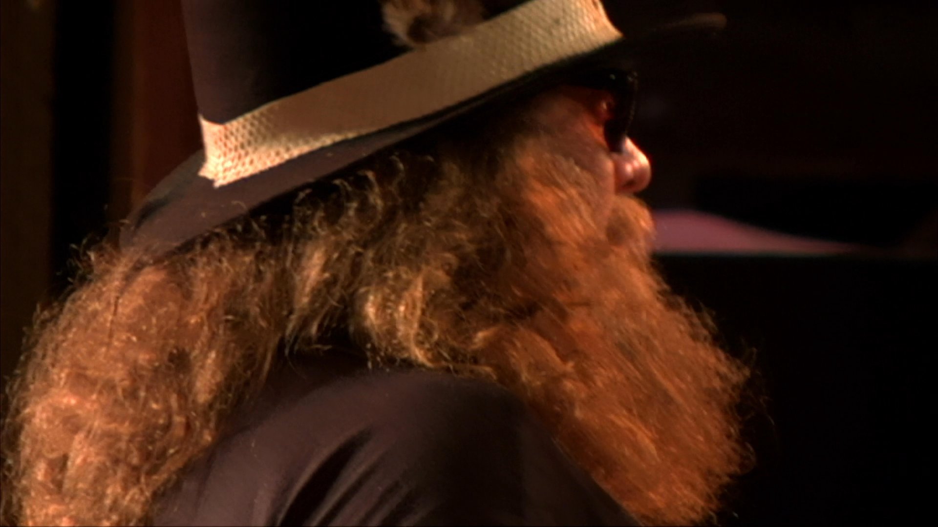 A long-haired, bearded musician performs live on stage with a fiddle and electric guitar while patrons dance in the background at a bar venue.