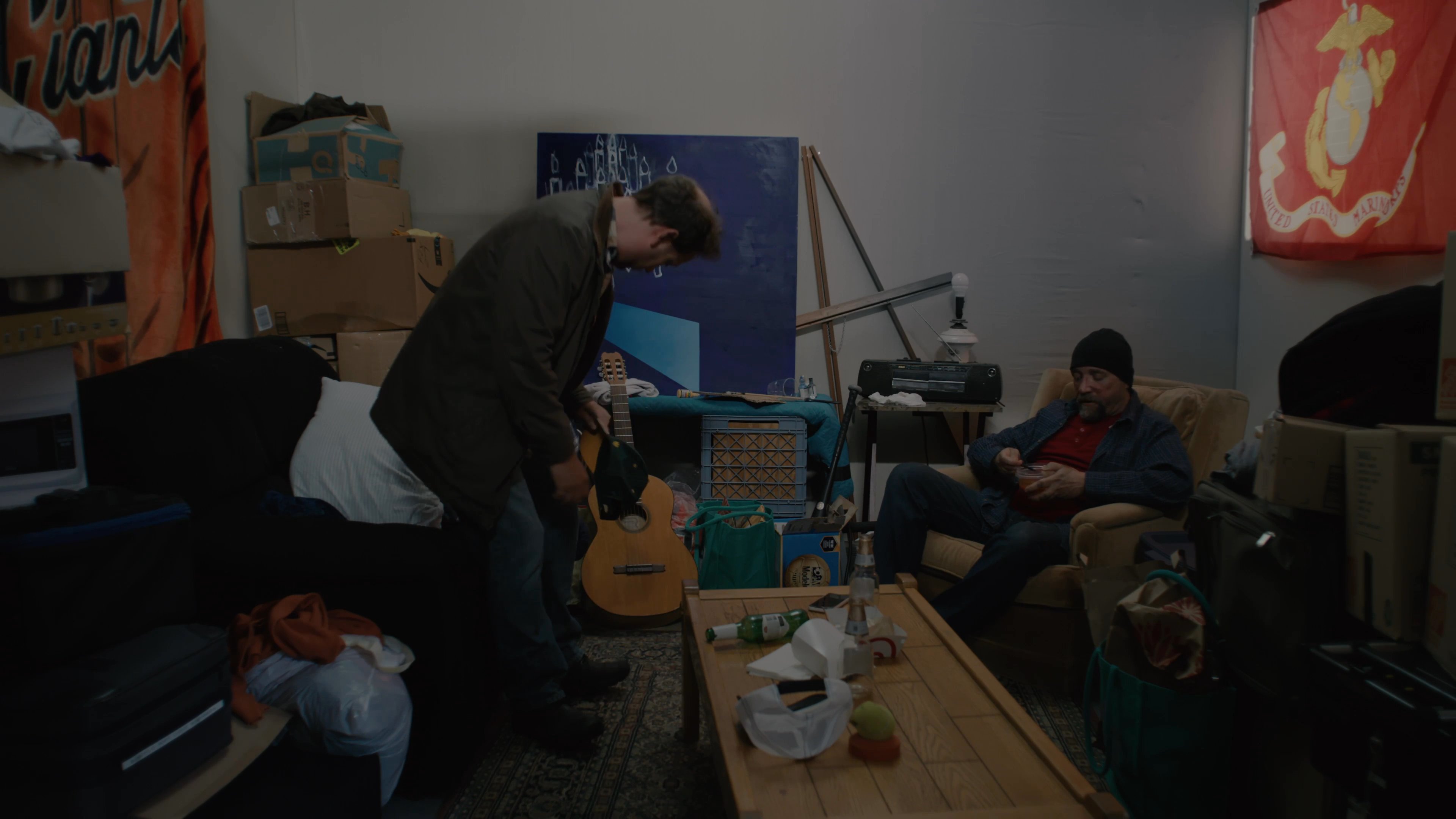 Two individuals engage in a relaxed dialogue while in a living room, with one person offering food to the other who is seated in an armchair.