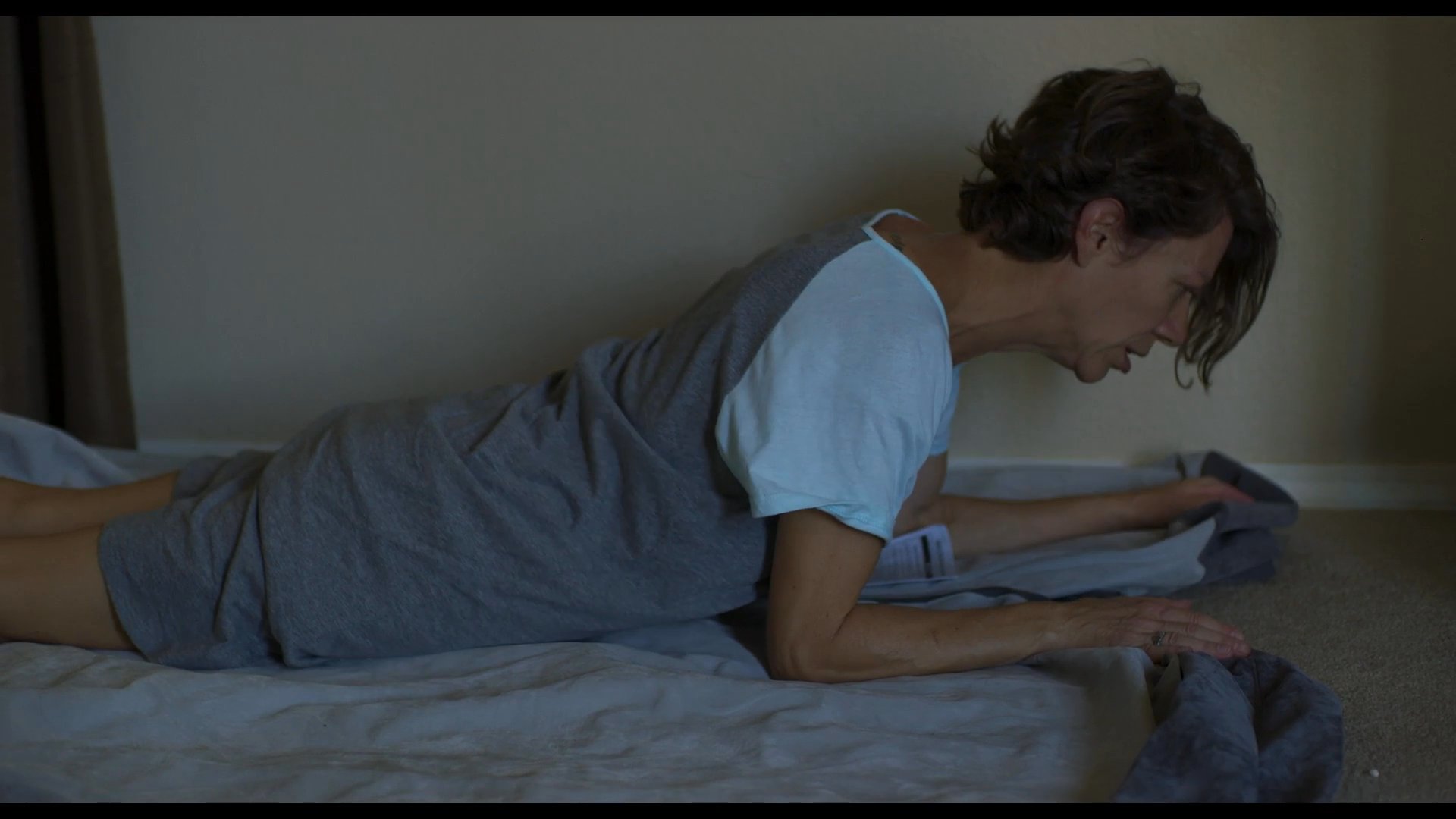 A woman exits a vehicle and crawls onto a bedroom floor, visibly distressed, before wrapping herself in a blanket to rest.