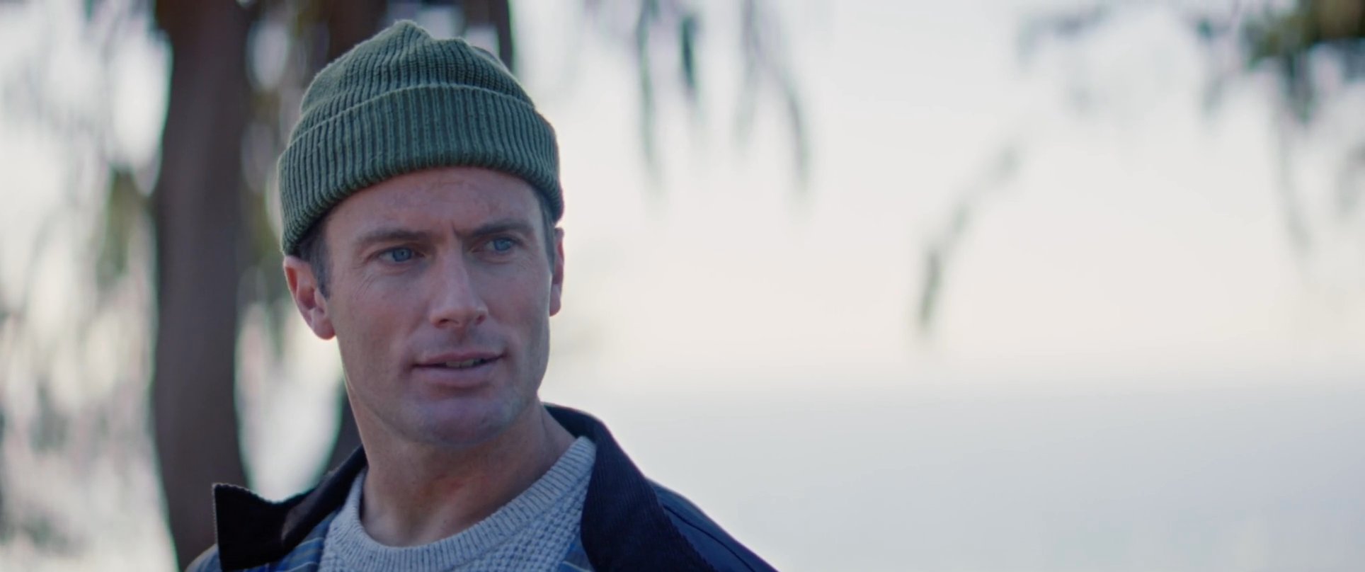 Two men in beanies engage in conversation while sitting on a park bench overlooking the ocean with their surfboards nearby. One man drinks from a metal mug as they share a quiet moment in a scenic coastal setting.