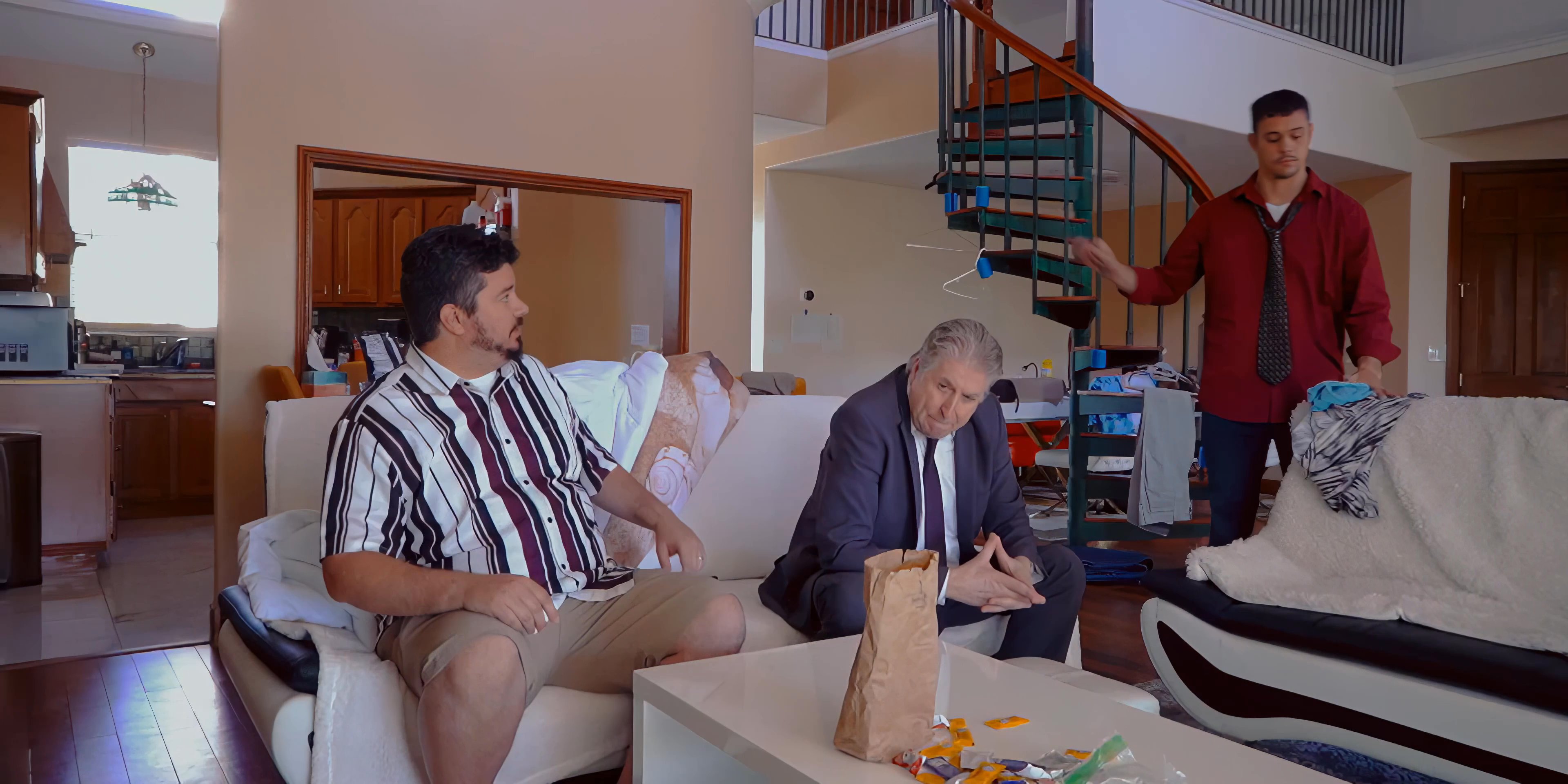 Three men engage in a tense, focused dialogue within a residential living area. The exchange involves movement and direct address, suggesting an emotionally charged or urgent professional interaction.