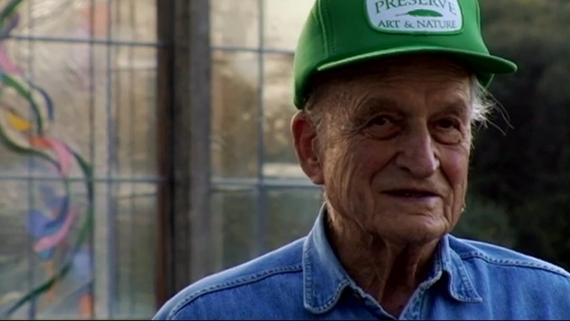 An elderly man in a green cap reflects on how personal taste shapes reactions to art. In a gallery or studio, several men handle and cover large paintings, including a vivid multicolored piece.