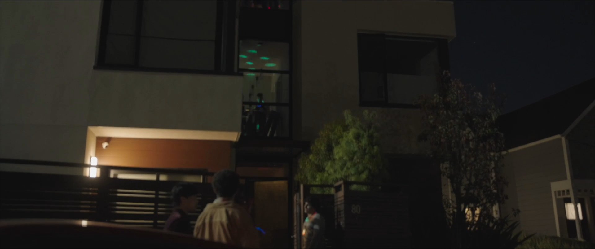 Two individuals arrive at a modern residence to join a social gathering, greeted by friends in an illuminated evening setting.