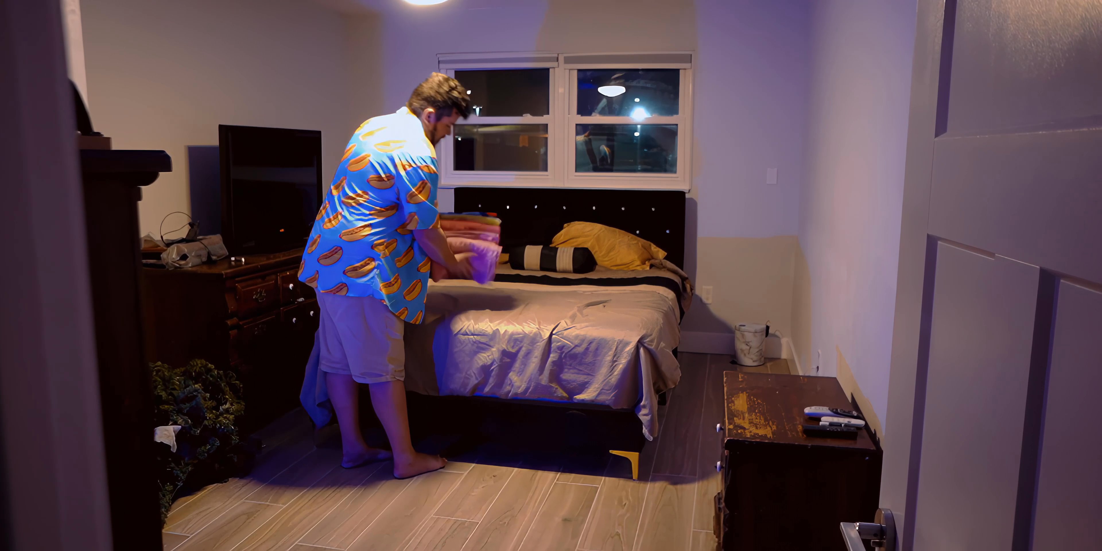 A man in a patterned shirt enters a bedroom at night and meticulously organizes blankets and bedding on the bed.