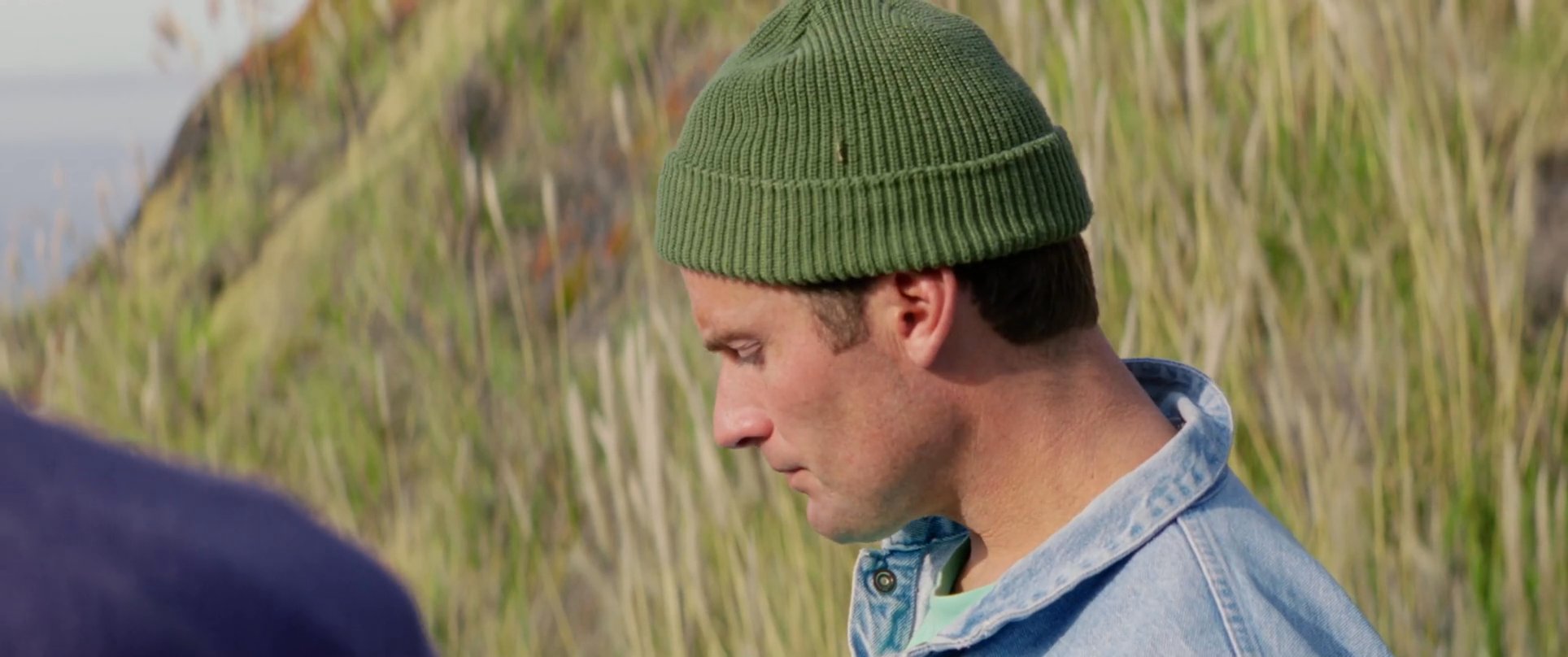 Two men stand on a grassy cliffside overlooking the ocean, engaged in an active, thoughtful conversation. The interaction transitions from a close-up dialogue to a wider shot of them walking together along the coastal path.