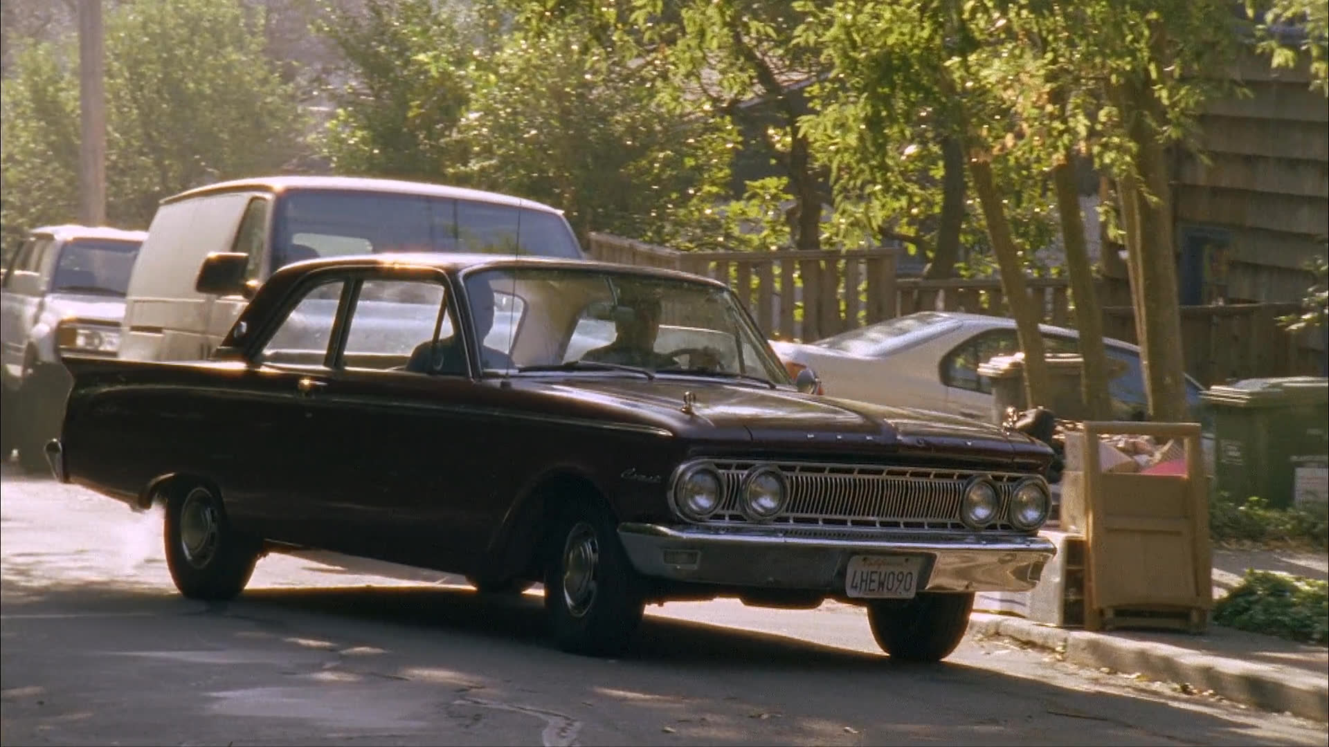 A classic dark-colored sedan drives along a residential street lined with parked vehicles and mature trees. The vehicle proceeds steadily past a white van as it moves down the road during the day.