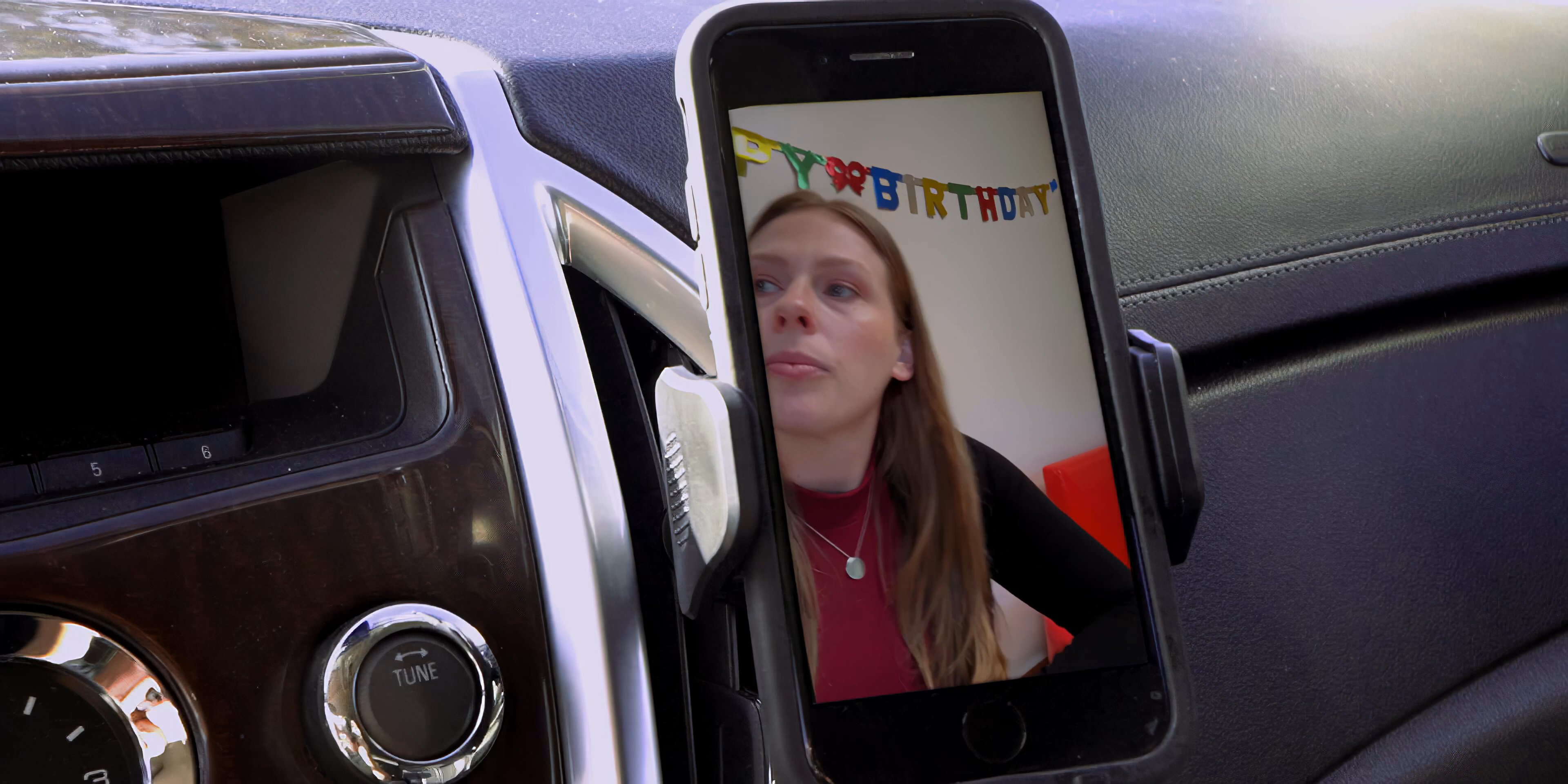 A man in a parked car engages in a remote video call, observing a screen showing a child and a woman in a celebratory setting.