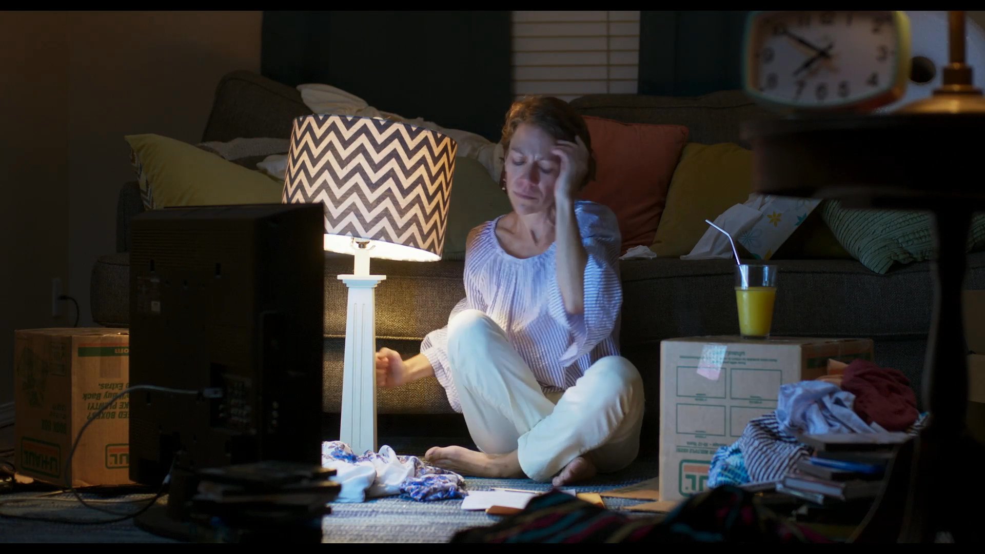 A woman sits on the floor of a dimly lit living room, maneuvering a lamp to provide better illumination while she reviews paperwork. She focuses intently on the documents amidst a setting of household clutter.