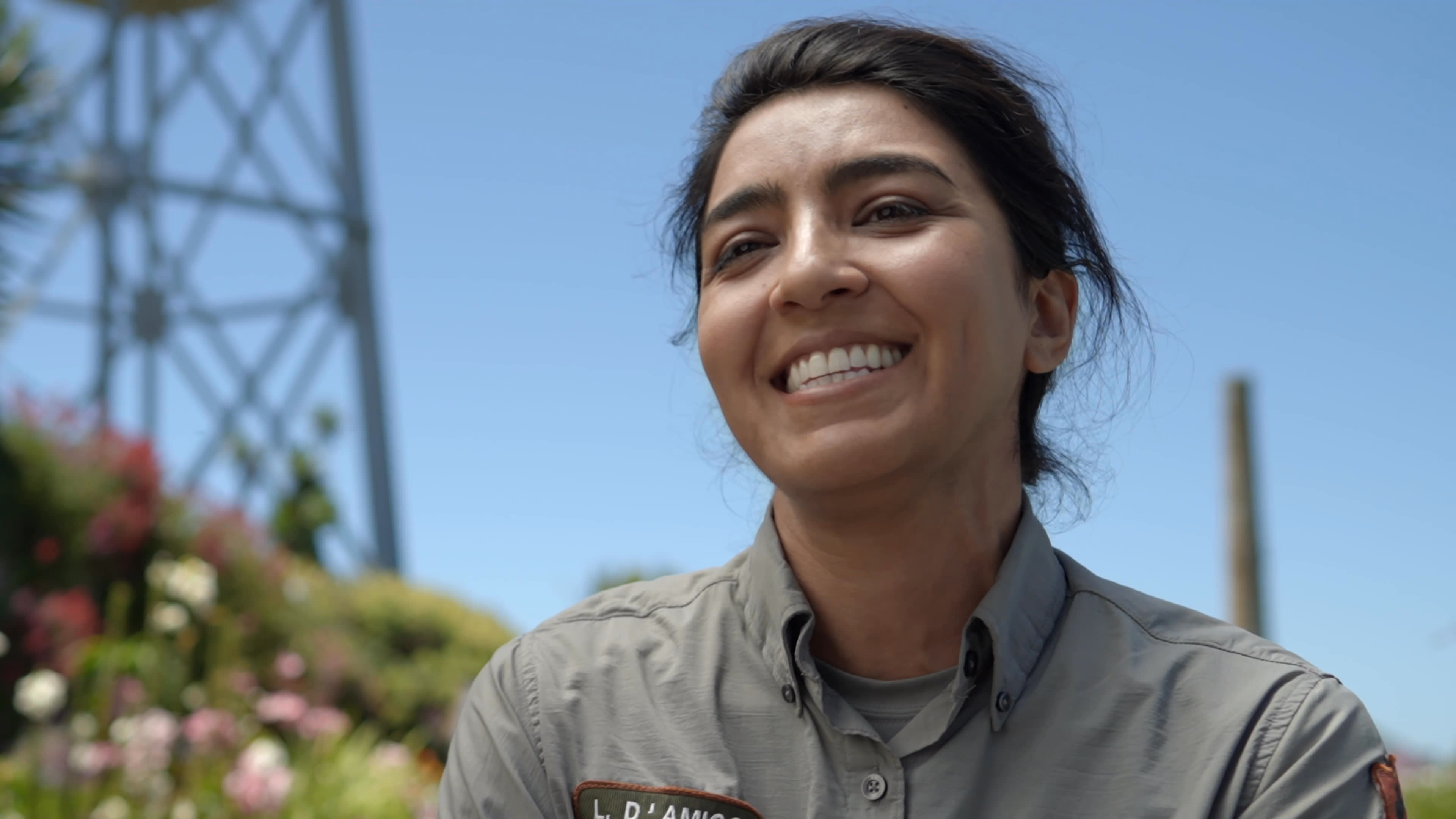 A wildlife professional speaks about her career outdoors beside a coastal garden, then walks with a companion as birds fly nearby. An adult falcon rests behind enclosure bars in close-up.