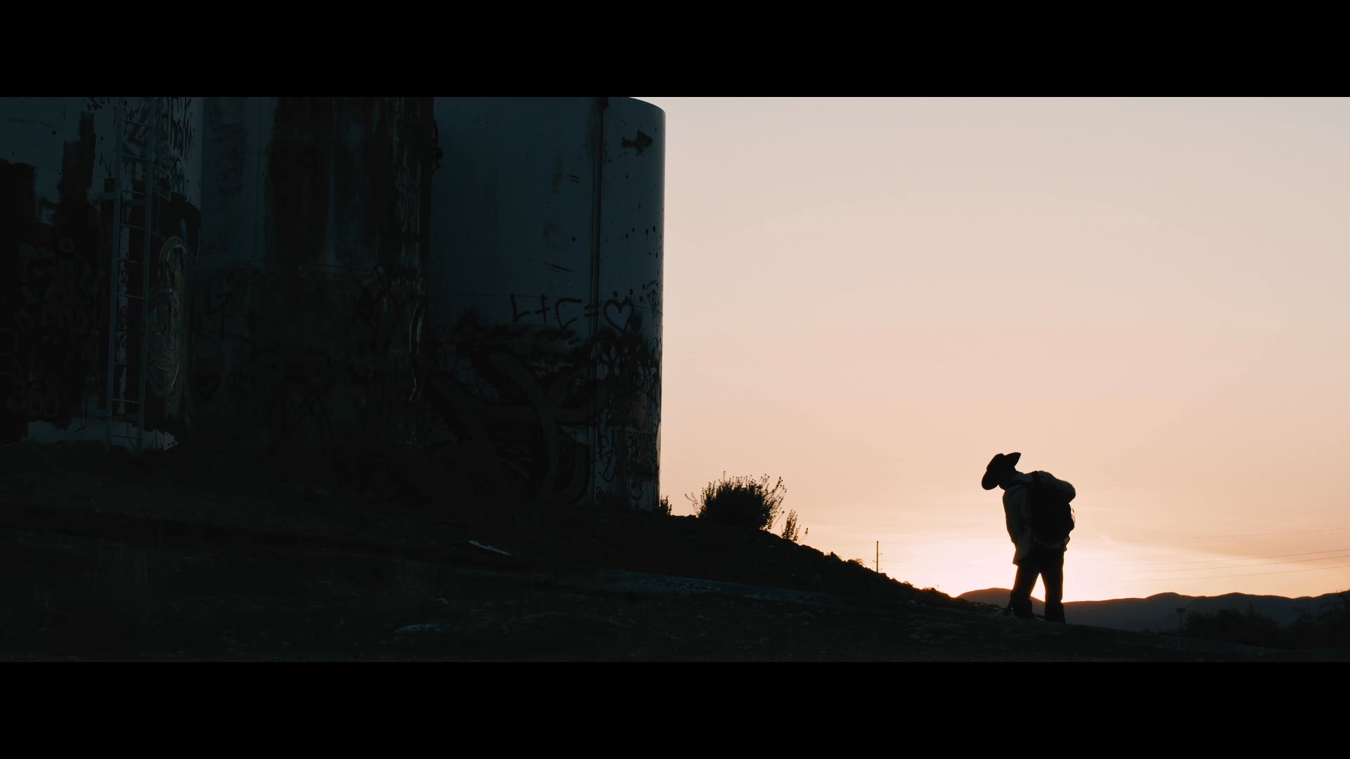 A silhouette of a worker digs in the desert at sunset near a large border wall. The scene captures the isolation and labor associated with the Mexico-USA border region.