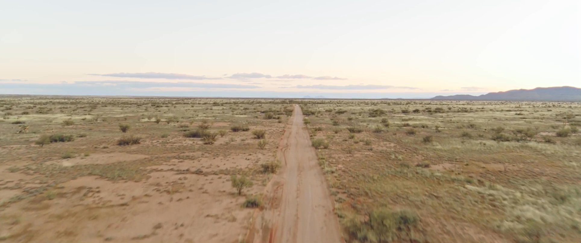 A lone traveler journeys across an expansive, arid desert landscape at daybreak. The scene captures the transition from a quiet campsite to a solitary trek along a remote dirt road.