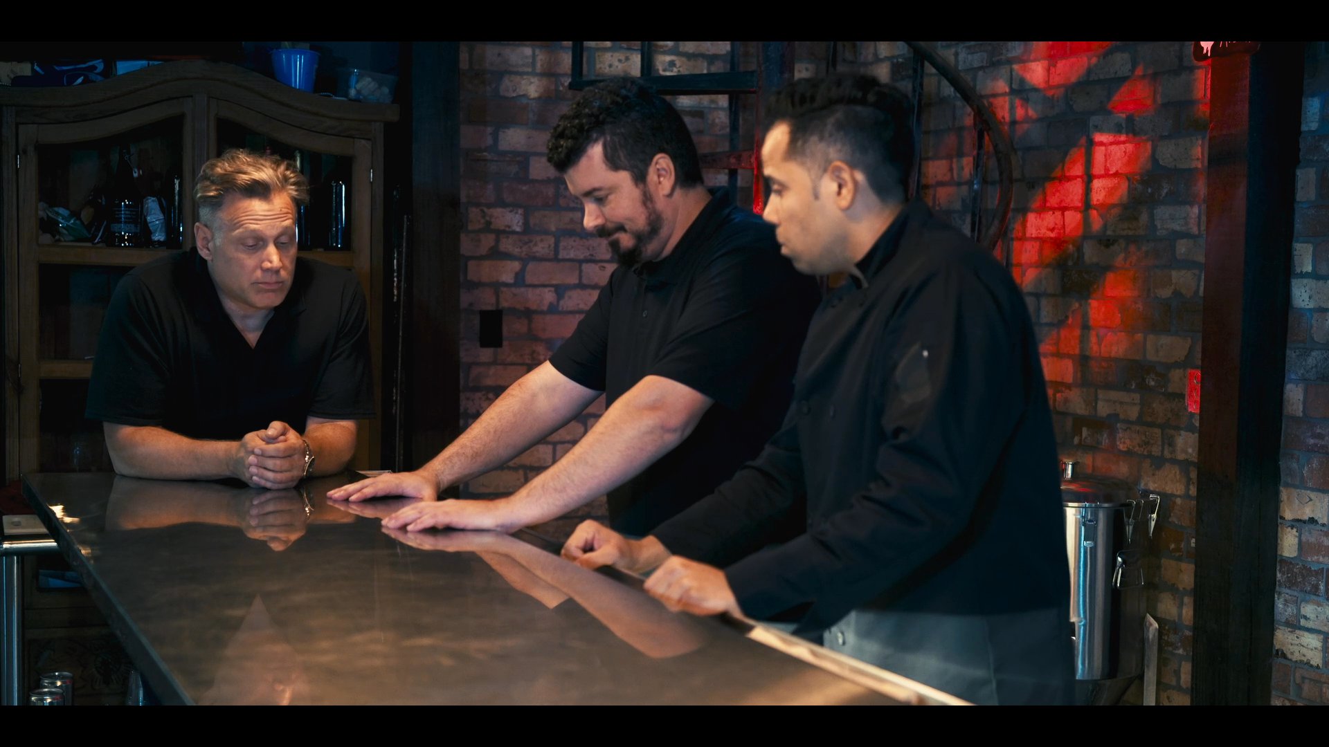 Three individuals engage in a focused discussion at a stainless steel counter within a professional kitchen environment. They exchange information and coordinate tasks in a collaborative setting.