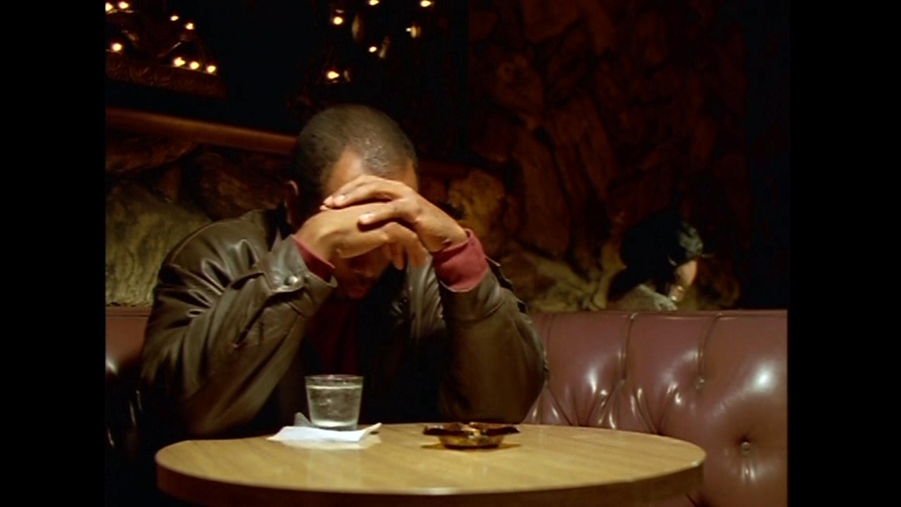 A man sits alone at a table in a dimly lit bar, displaying signs of deep contemplation or distress before lighting a candle. The sequence highlights a somber, intimate atmosphere.