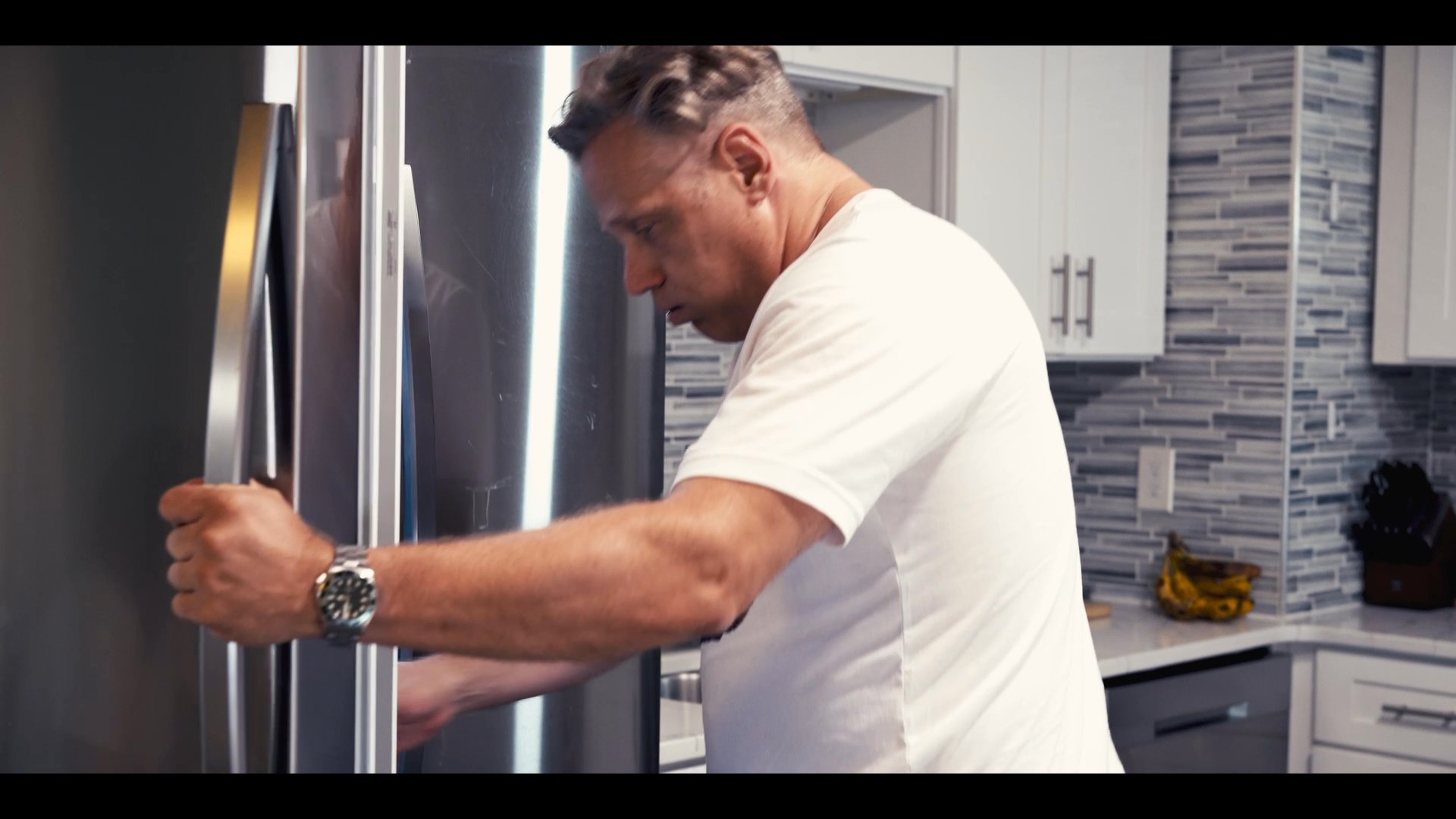 An aerial perspective transitions into an interior view of a man navigating a modern kitchen to retrieve a beverage from the refrigerator. He consumes the drink before returning the container to the kitchen area.