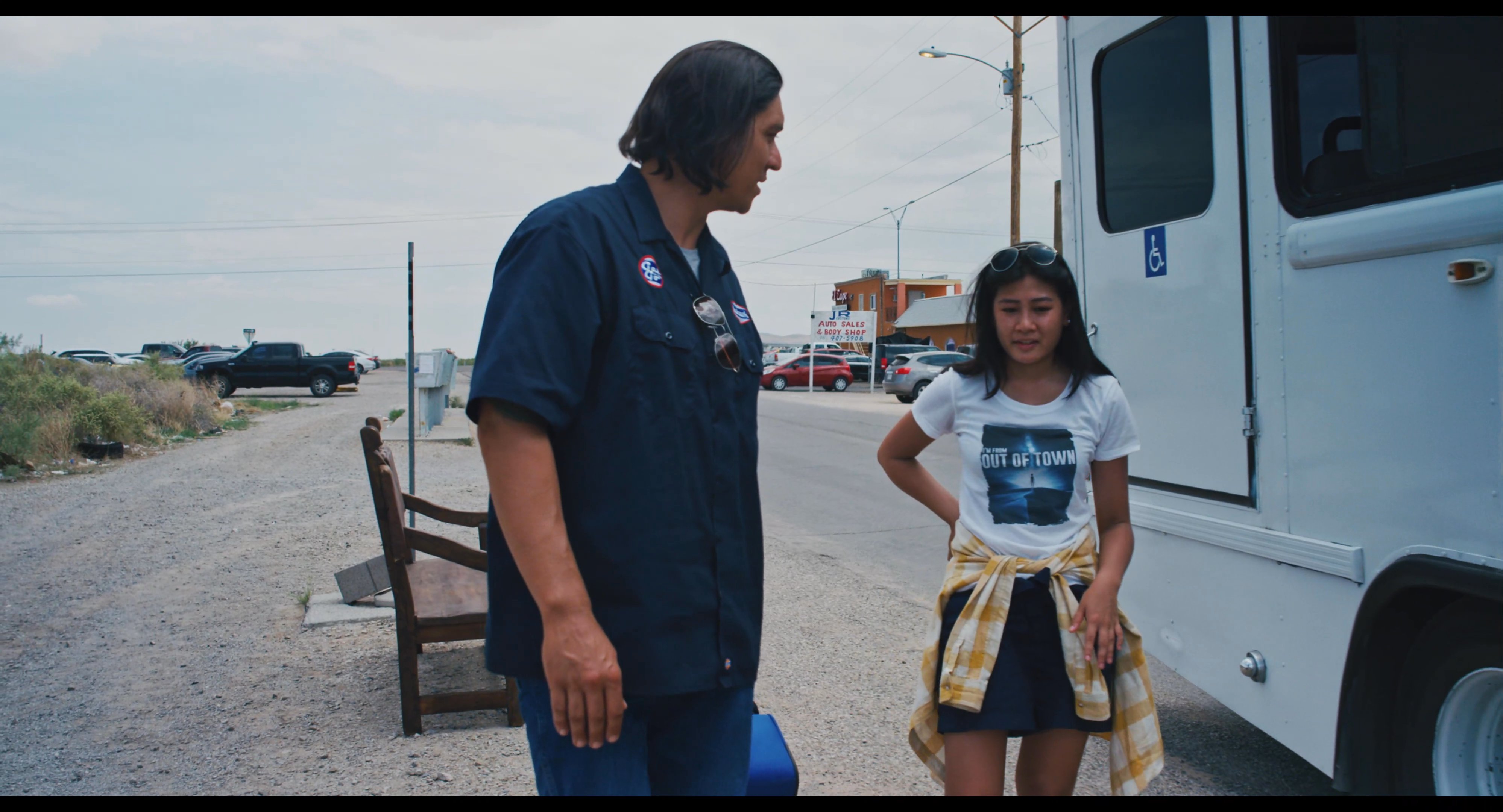 A man and a young woman engage in a final conversation beside a parked bus while holding luggage. They exchange words before the woman prepares to board the vehicle.