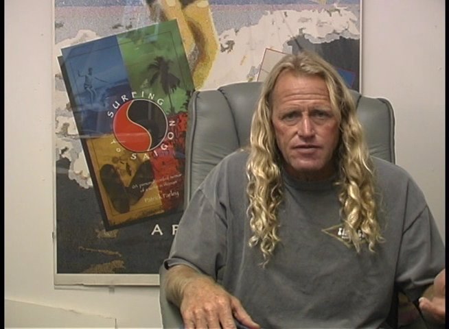 A man with long blonde hair discusses surfing while sitting in a professional office environment, using hand gestures to emphasize his points.