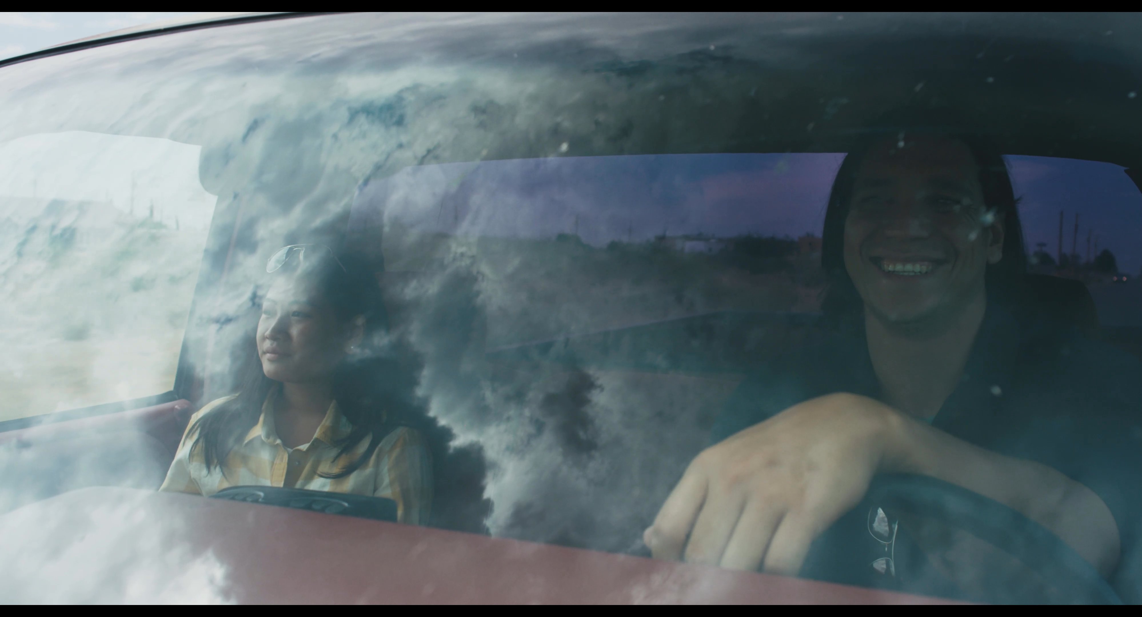 Two individuals sit inside a car parked before an alien-themed storefront in the desert, reflecting on their journey under a cloudy sky.